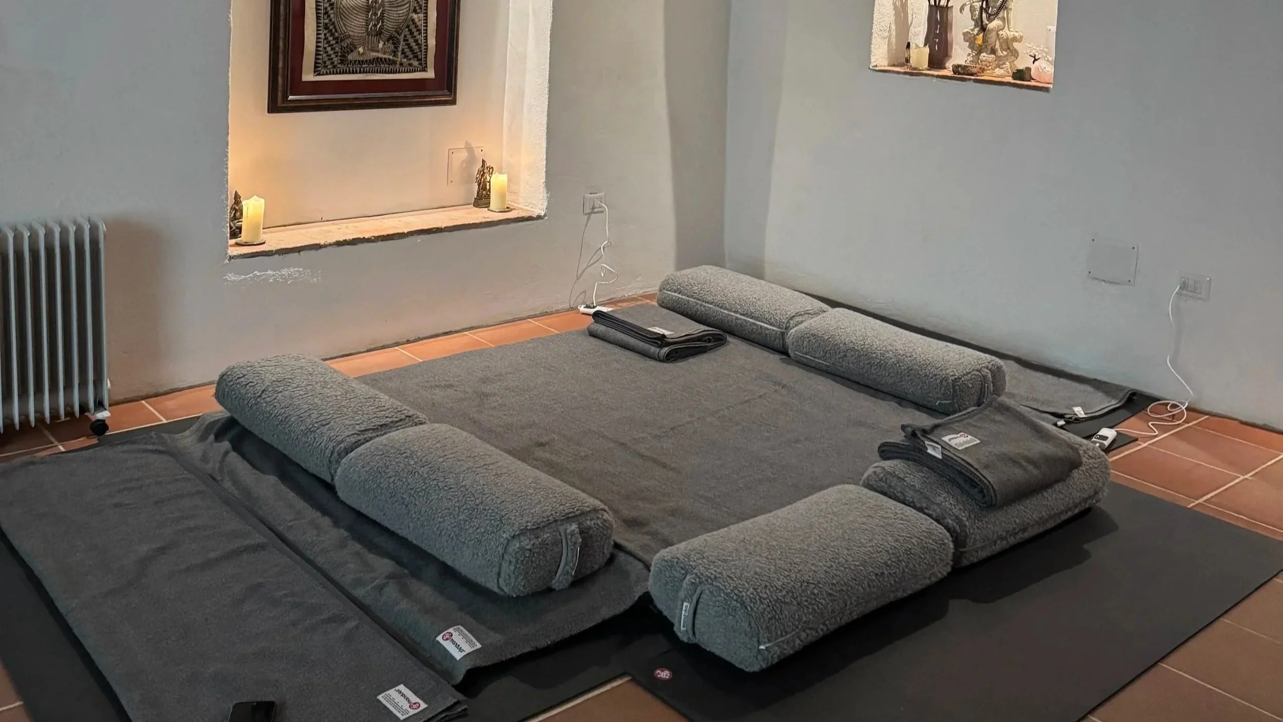 A living room with a low, gray cushioned surface, possibly a bed or seating, surrounded by rolled towels or cushions. There are candles, small decorative items, and artwork on the walls. An electrical cord and outlet are visible, with a vent radiator to the left and a small window on the right.