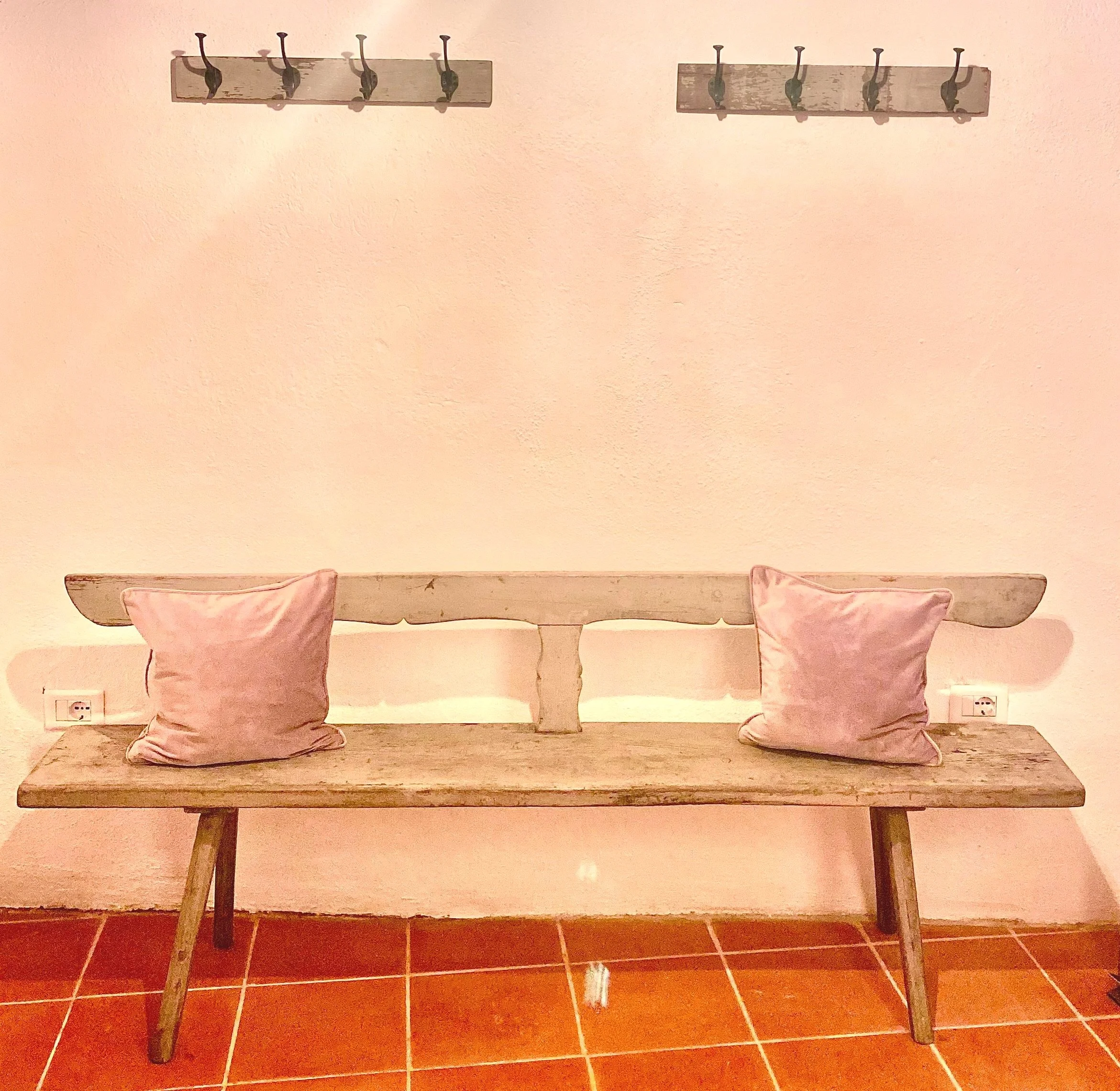 Wooden bench with two pink cushions, against a pink wall with two wooden coat racks above. The bench is on a red tile floor.