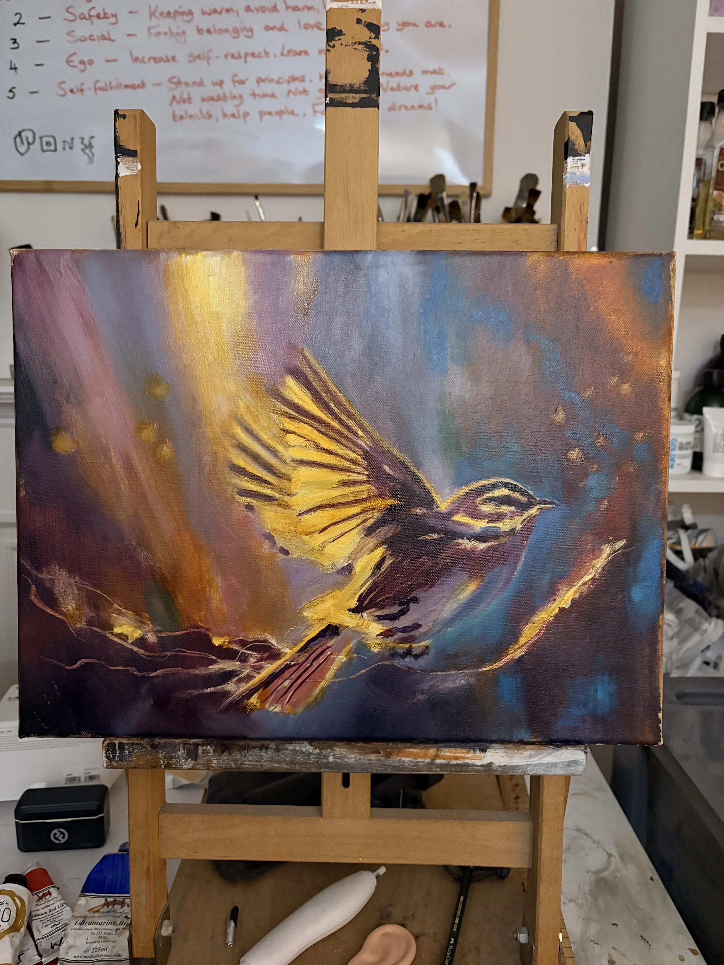 An oil painting of a bird soaring with outstretched wings, painted with bright yellow, purple, and orange hues, on an easel in a studio.
