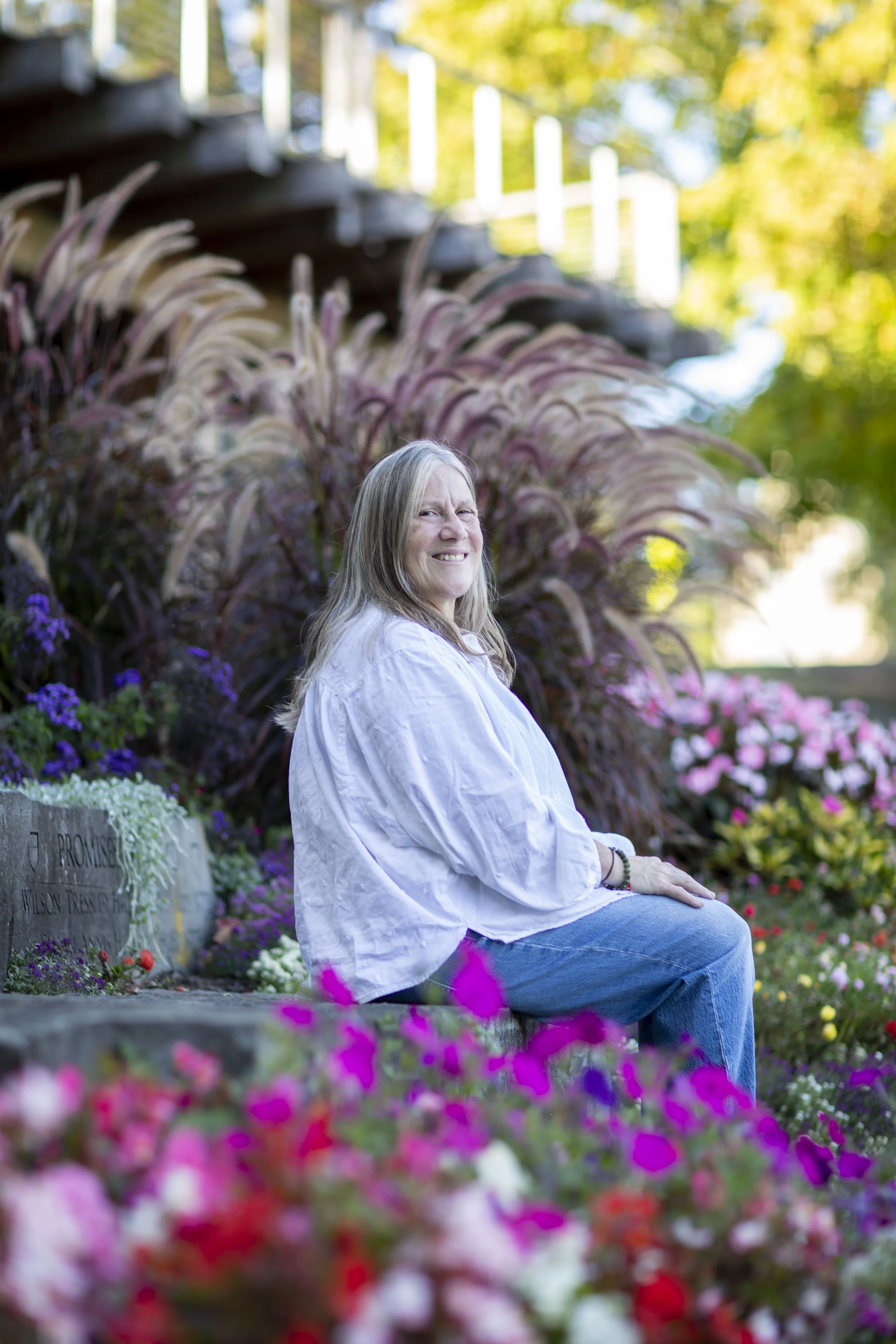 A woman with long gray hair sitting outdoors on a stone ledge surrounded by colorful flowers, with trees and a staircase in the background.