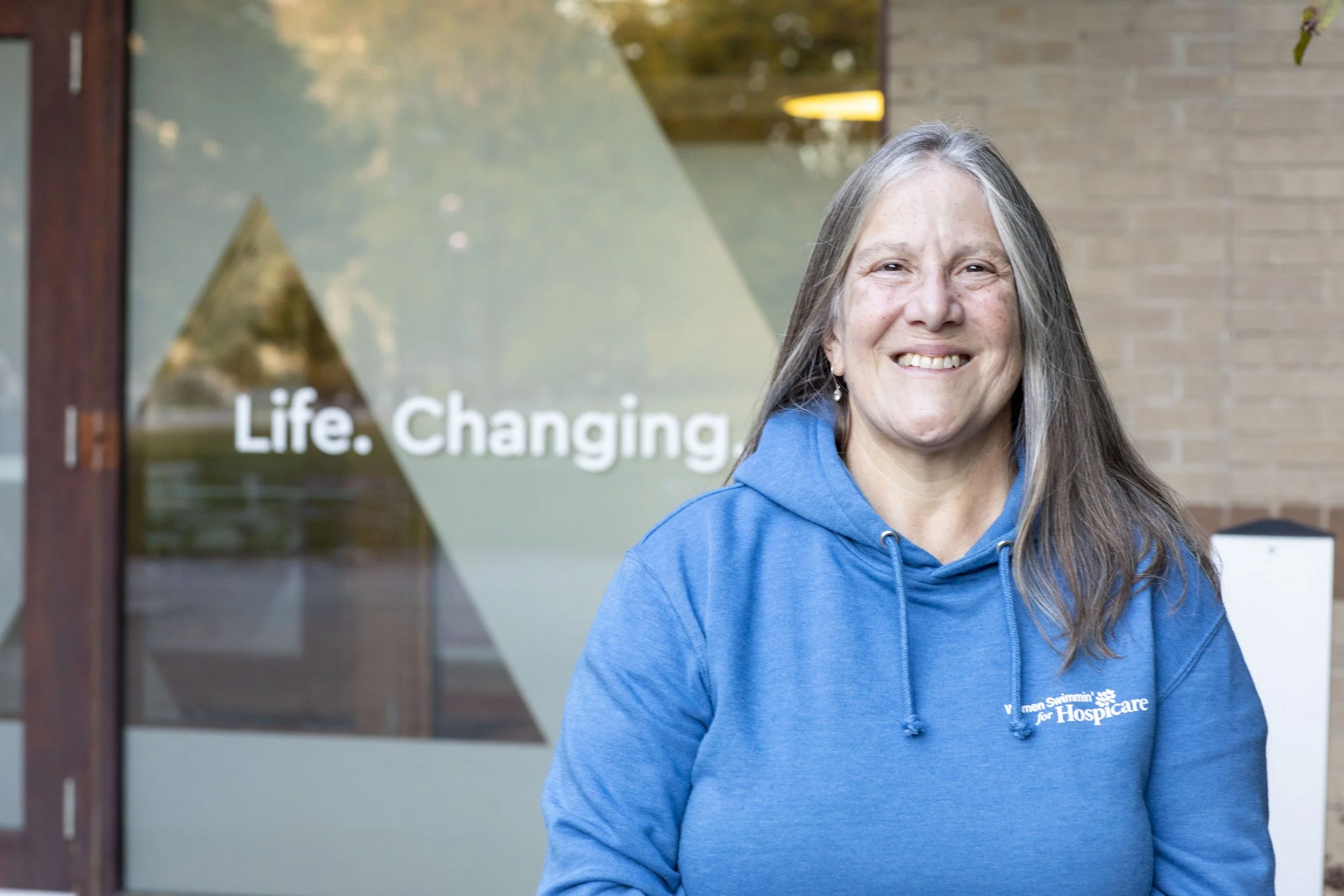 A smiling woman with long gray hair wearing a blue hoodie stands outdoors in front of a building with a glass door that has the words 'Life. Changing' written on it.