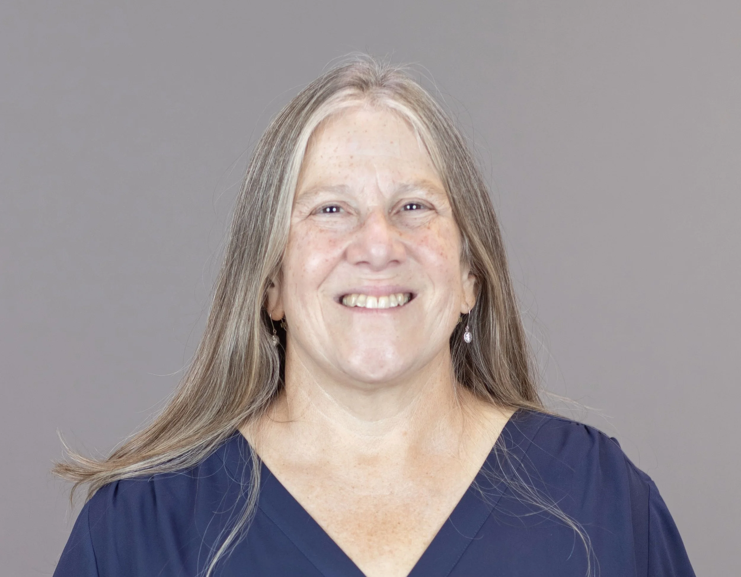 A smiling woman with long gray hair wearing a navy blue top and earrings, standing against a gray background.