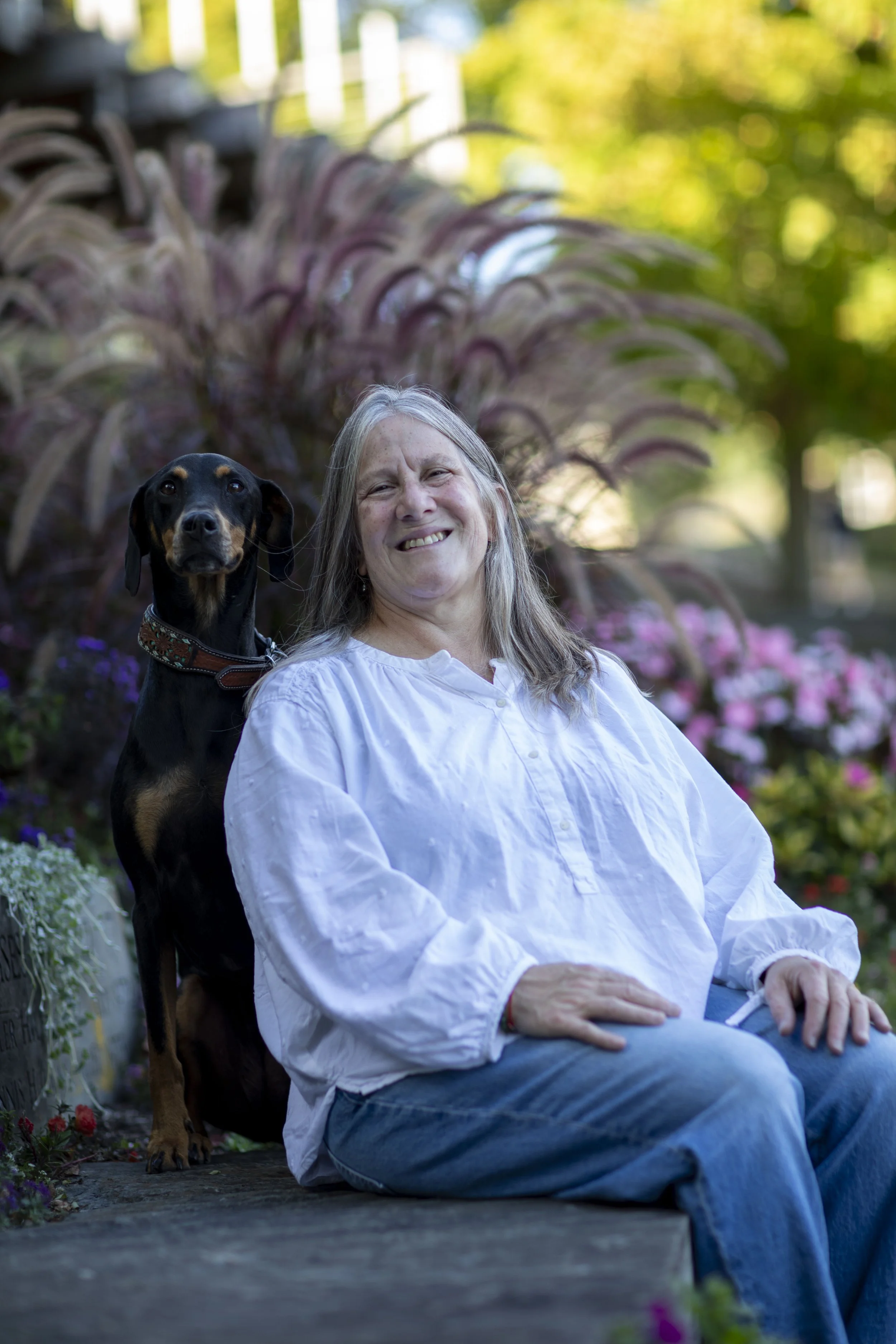 A smiling woman sitting outdoors in a garden with her dog standing beside her. The woman has long gray hair and is wearing a white shirt and jeans. The garden features green trees and colorful flowers, with sunlight filtering through the foliage.