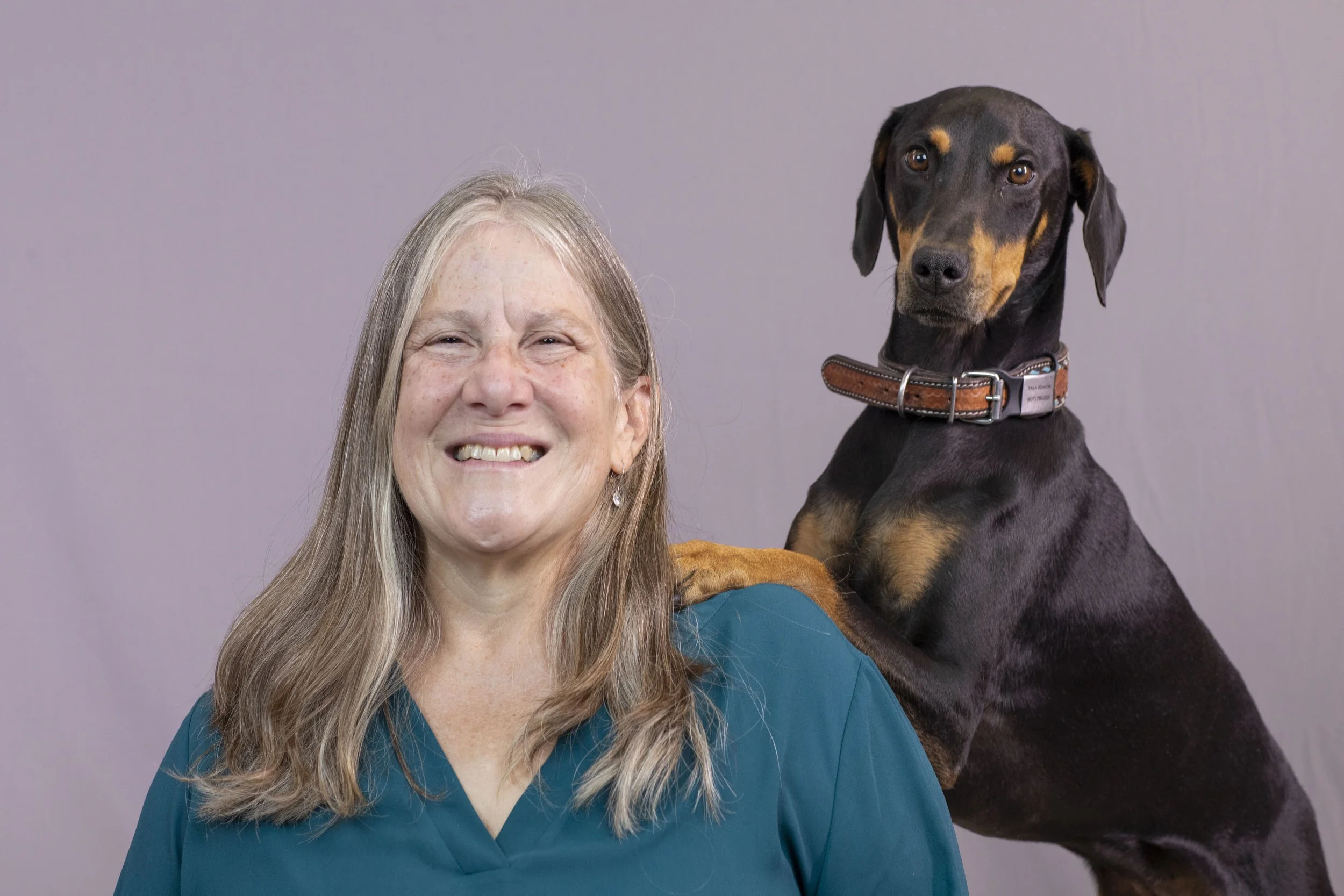 A smiling elderly woman in teal scrubs with a Doberman pinscher dog standing behind her, resting its paw on her shoulder, against a plain gray background.