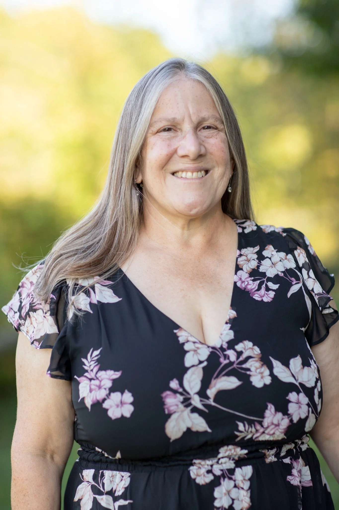 A woman with long, gray hair smiling outdoors against a blurred background of green and yellow trees, wearing a black dress with pink and white floral pattern and pearl earrings.