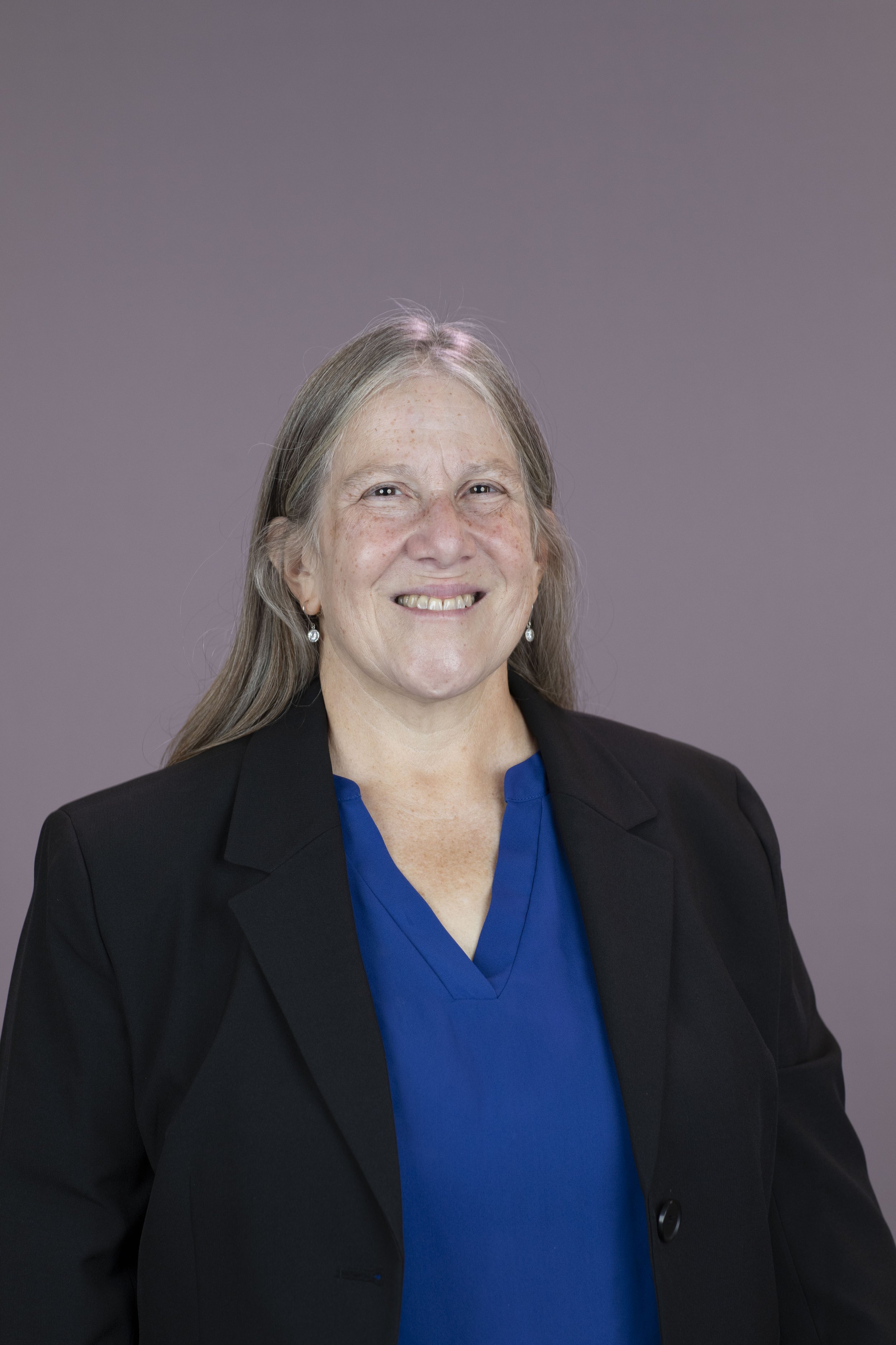 Portrait of a smiling middle-aged woman with gray hair, wearing a black blazer over a blue top, standing against a plain gray background.