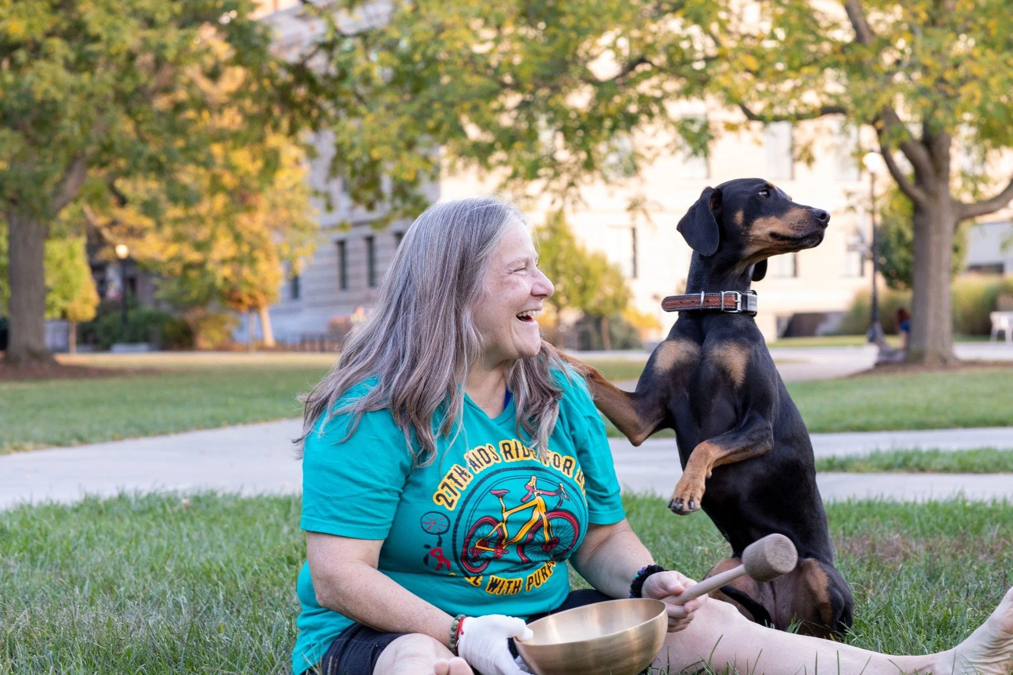 A woman with long brown hair sitting on grass at a university, holding a sound healing bowl, smiling at a black and tan mixed breed dog that is sitting on its hind legs, with one paw on her shoulder. The background shows trees and a building.