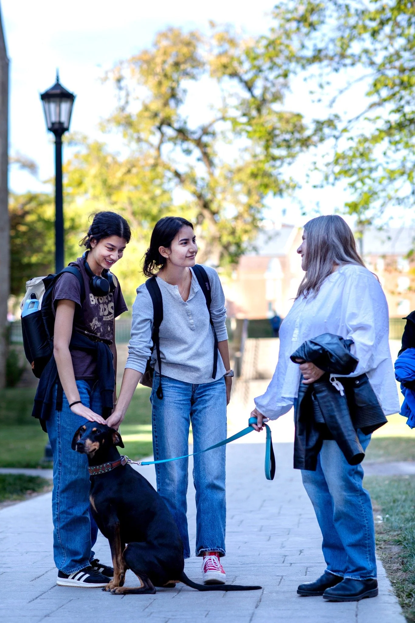 Two young women with backpacks talking to an older owman with brown hair on a sidewalk at a university with trees and a lamppost in the background, accompanied by a black and tan dog on a leash.