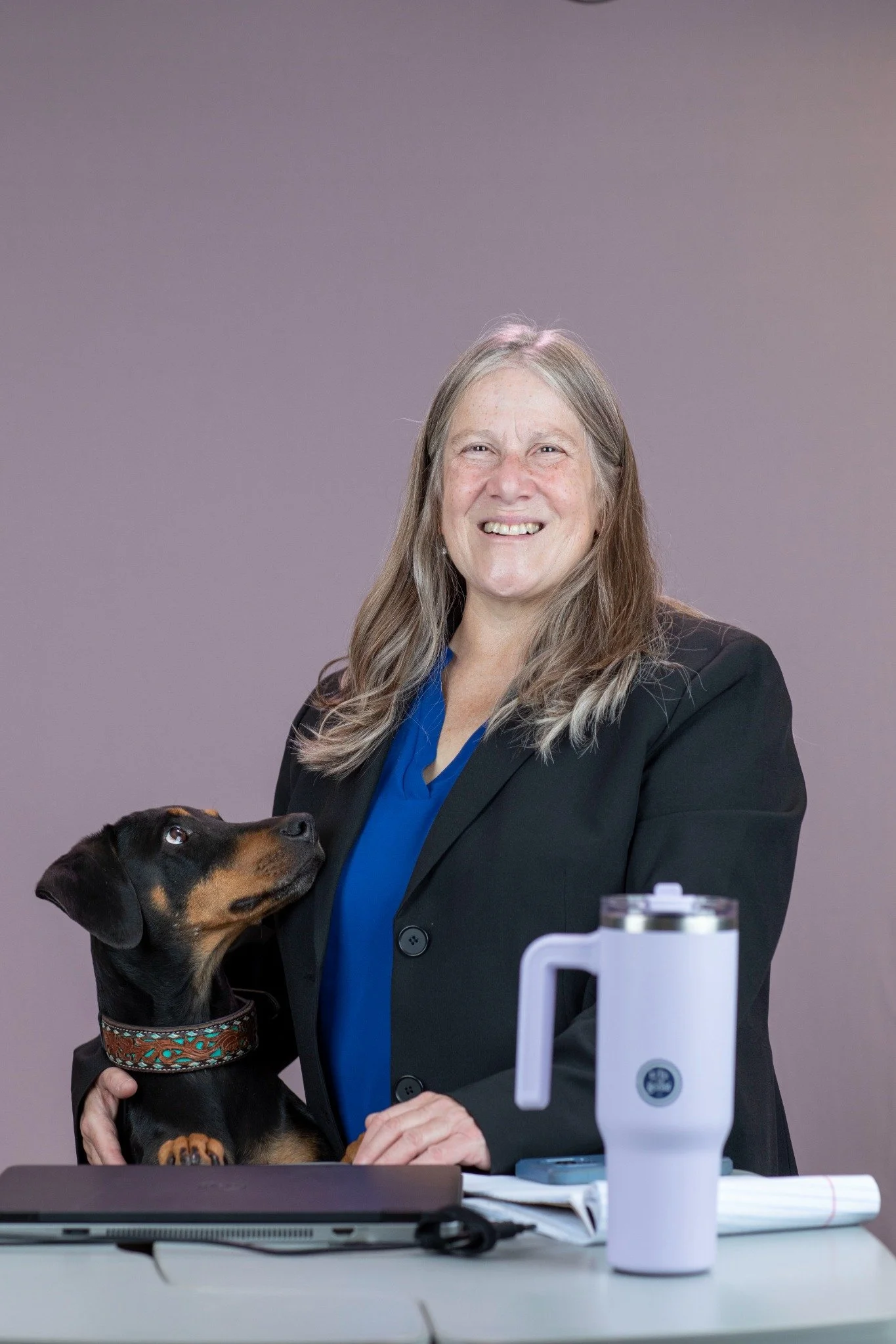 A long brown haired smiling woman wearing a black blazer and teal shirt at a table with a laptop, a tumbler with a yoga image, and papers. A black and tan mixed breed dog with a colorful collar stands upright next to her The background is plain gray.
