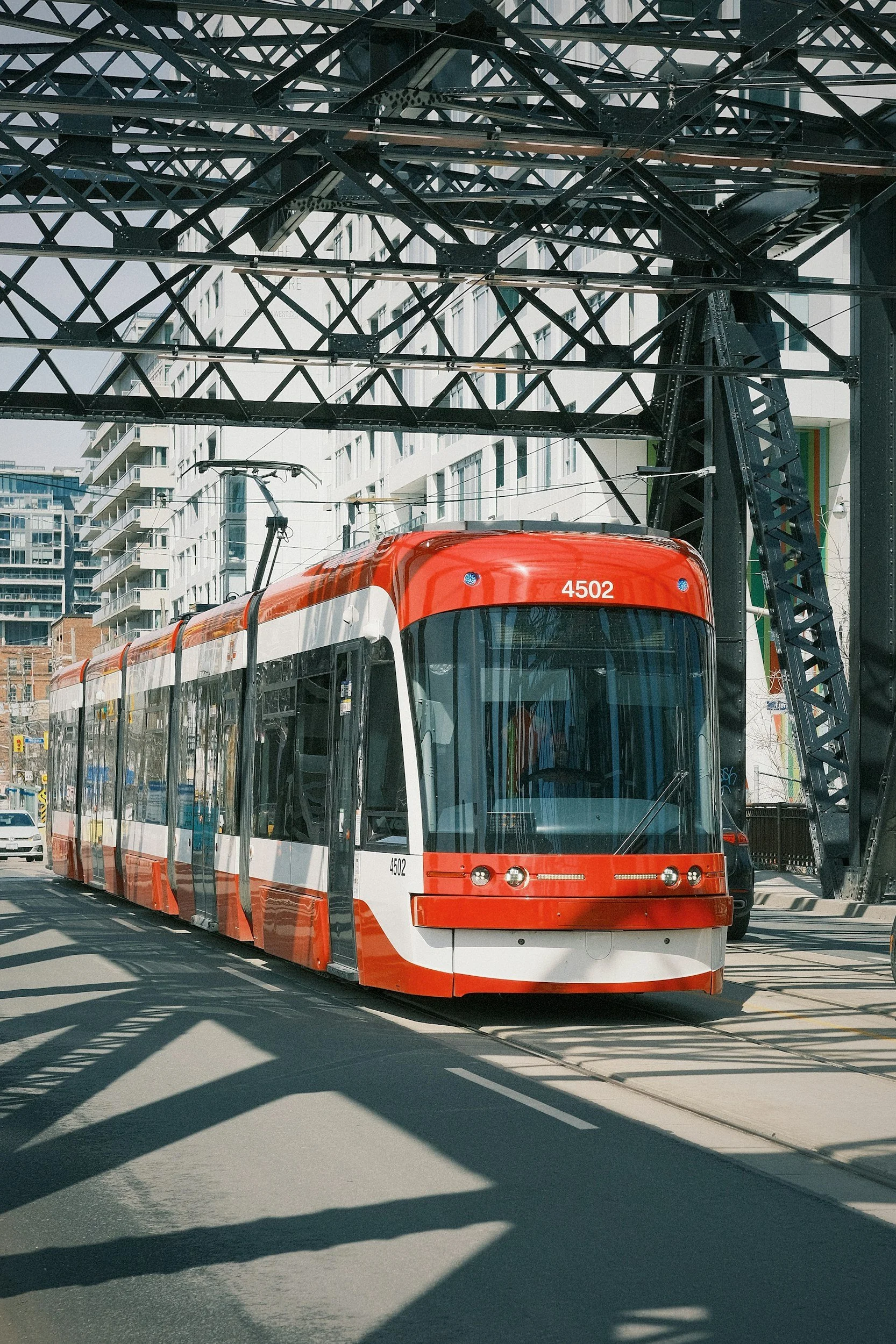 A modern red and white streetcar on tracks beneath a metal bridge in an urban area with residential and commercial buildings in the background.