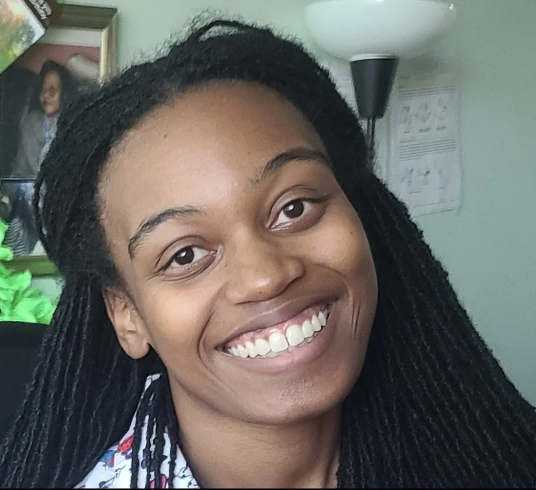 Close-up of a smiling woman with black dreadlocks, wearing a gray jacket with gold buttons.