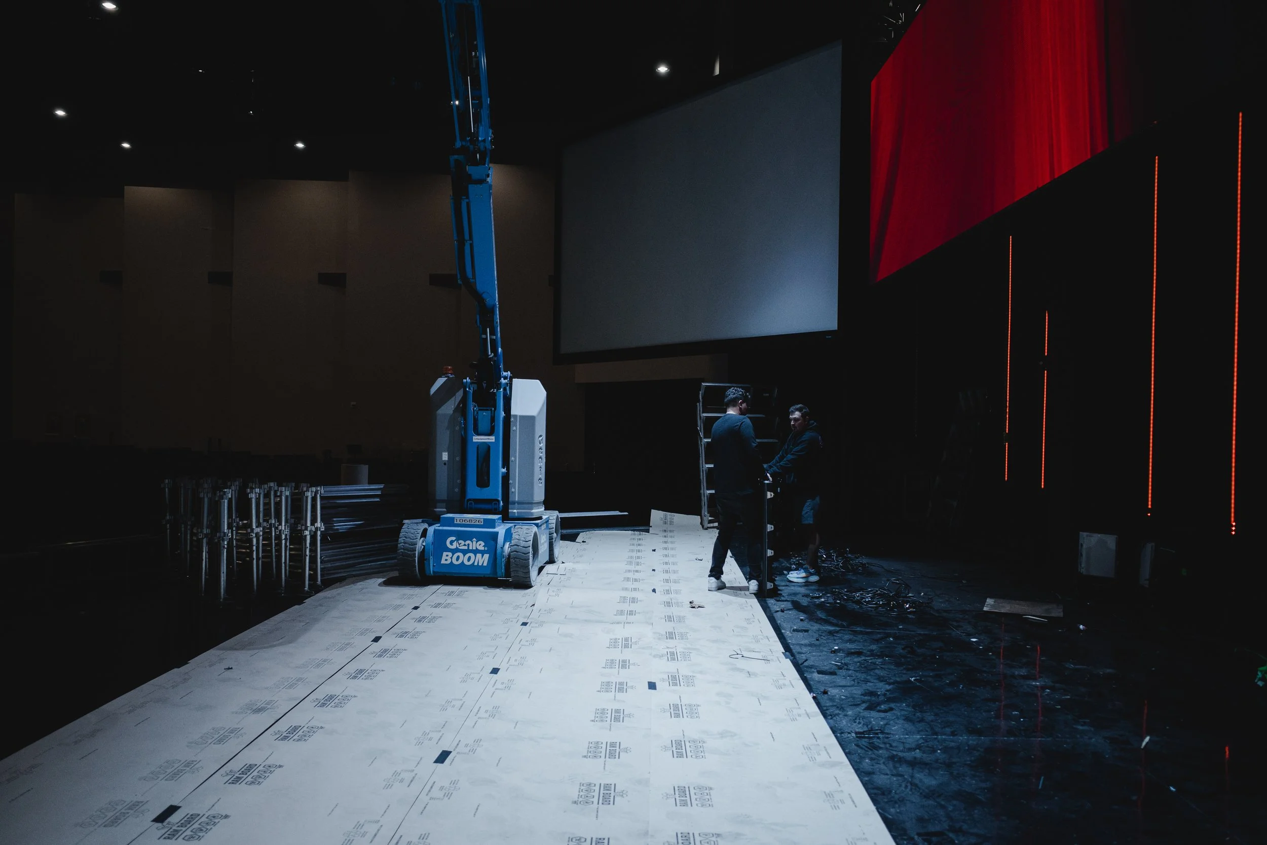 Under construction or setup stage in a theater or auditorium; with two people talking near a projector lift, a large screen, and on the left, materials and equipment on the stage.
