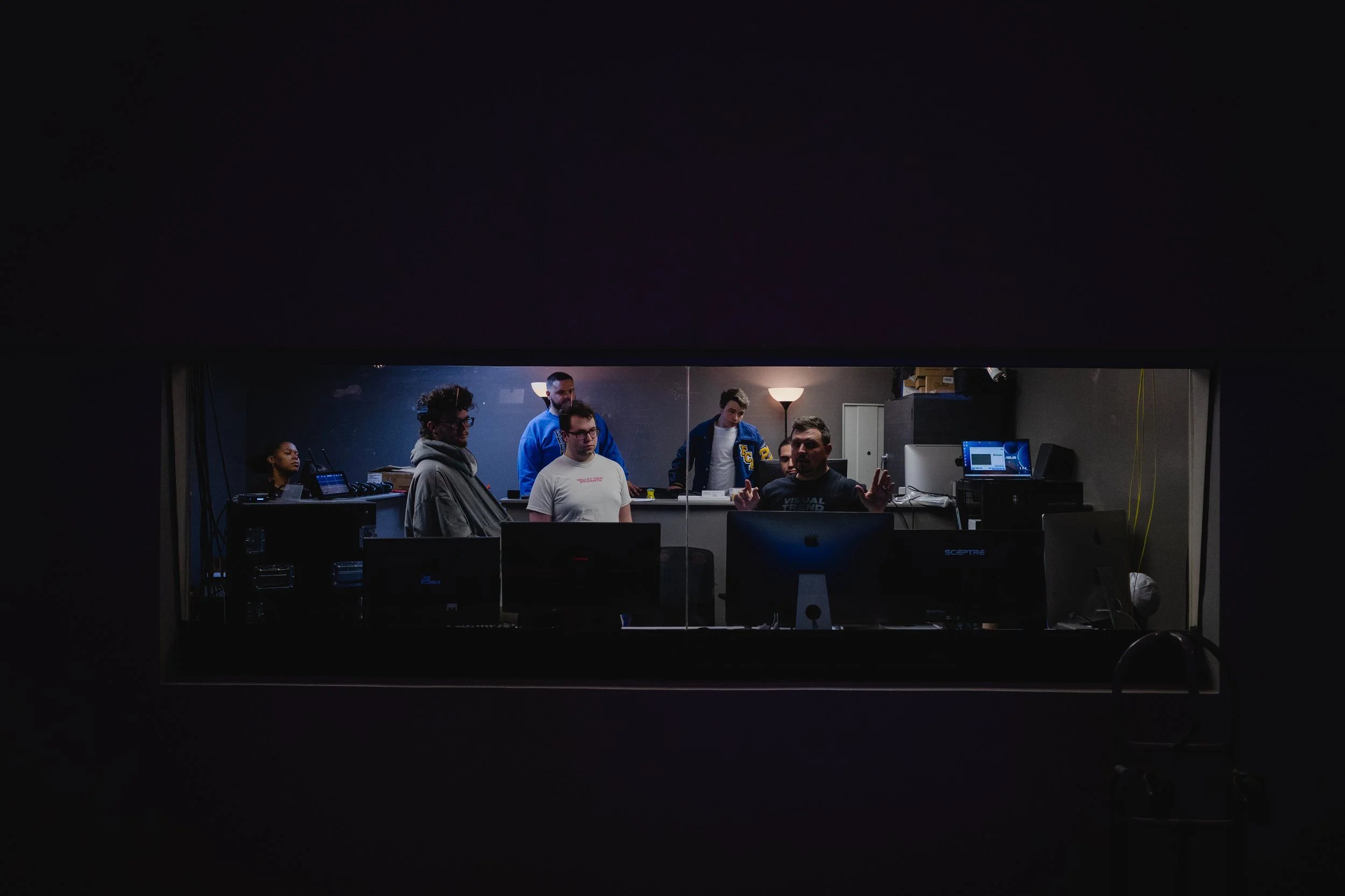 View through a window of a church tech team of training together in the tech room or booth of the church, with various computers and equipment.