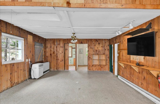 Empty living room with wood-paneled walls, a wall-mounted flat-screen TV, a window, a ceiling fan, and carpeted floor.