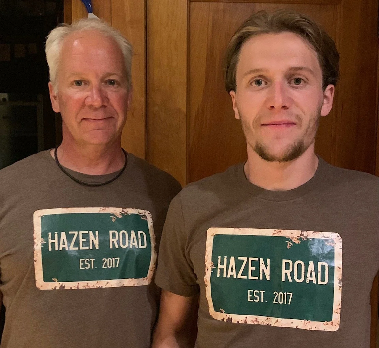 Two men standing side by side wearing gray T-shirts with a green and white road sign design that reads "Hazen Road EST. 2017." They are in a room with wooden paneled walls, and both are smiling.
