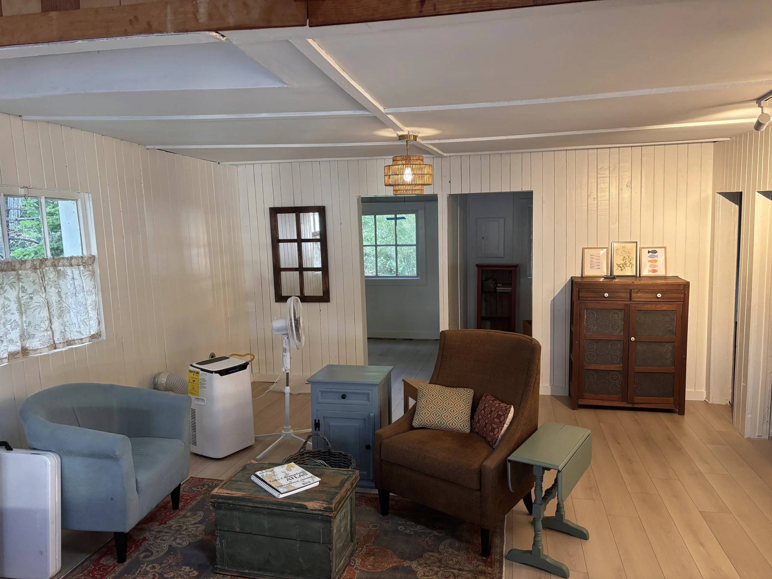 A cozy living room with white-paneled walls, hardwood flooring, and a window with floral curtains. Contains a brown armchair with pillows, a small blue side table, a wooden coffee table, a white fan, and a blue sideboard. There is a modern ceiling light fixture and a framed artwork on the wall.