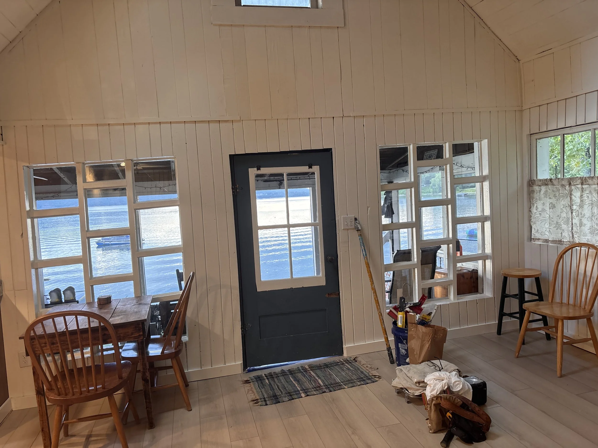 Interior view of a room with cream-colored wooden walls and large windows overlooking a body of water. The room contains a wooden dining table with chairs, a closed door, and various construction or renovation tools and materials including a broom, a bucket, and some cloth on the floor.