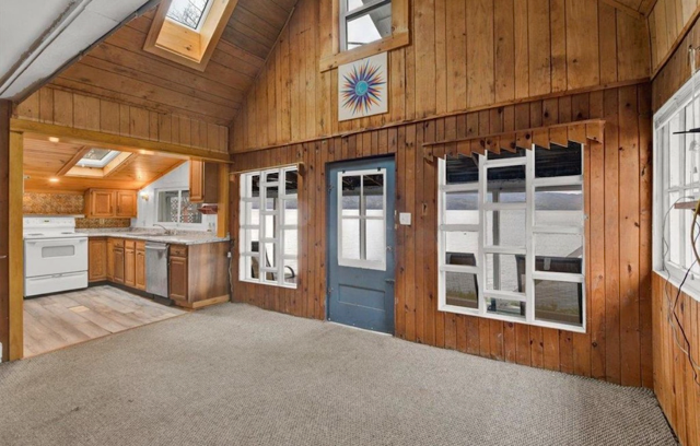 Interior of a wooden cabin room with a carpeted floor, windows, a blue door, and a view into the kitchen area with wooden cabinets and a white stove.