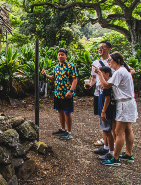 Four people, including a young boy, a woman, and a man, standing outdoors in a lush green park or garden, looking at an informational sign, with trees and plants in the background.