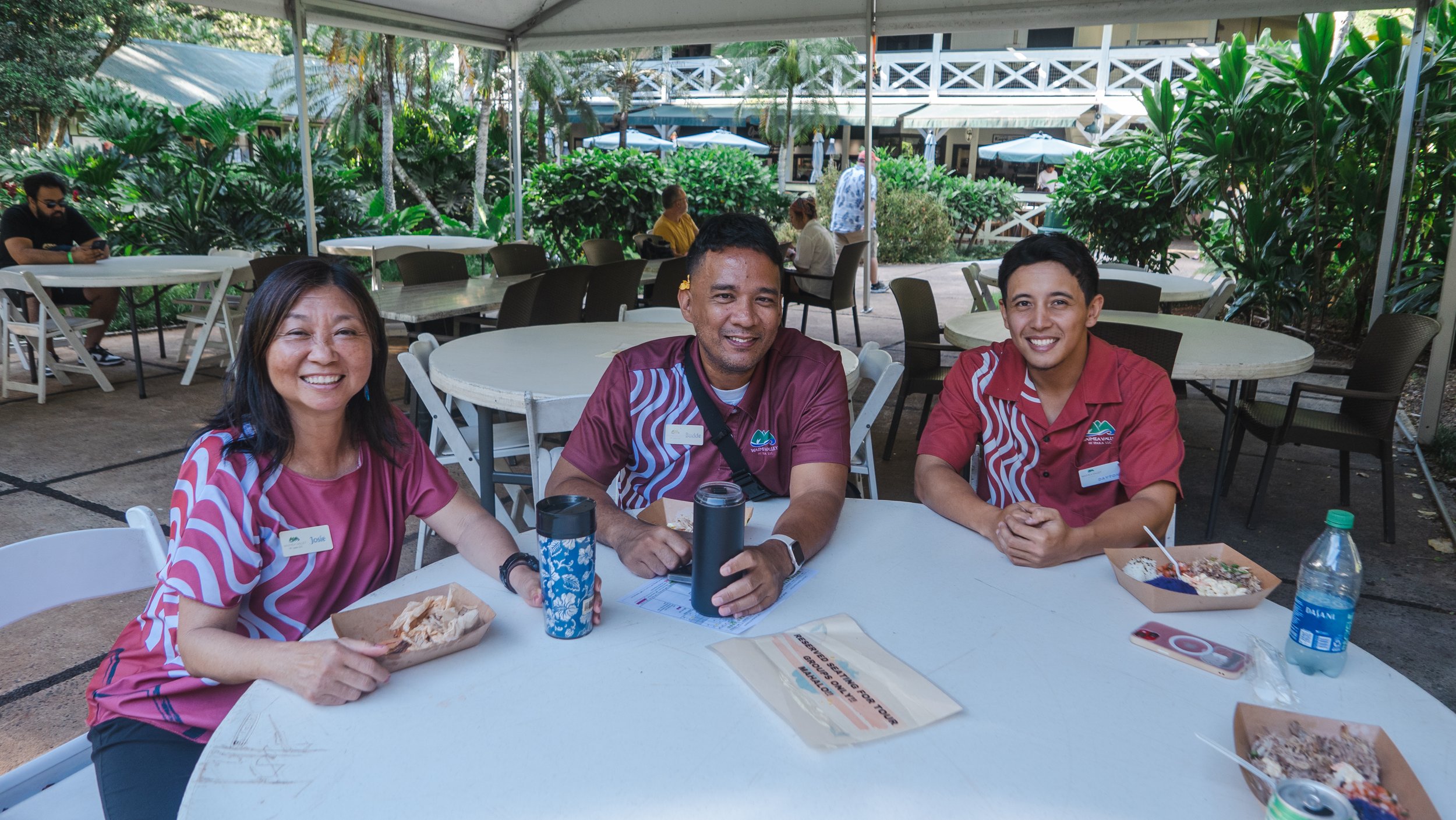 Three people sitting at a round table with food and drinks outdoors, smiling at the camera. Two women and one man, all wearing matching maroon shirts with a floral pattern and name tags, in a lush tropical setting.