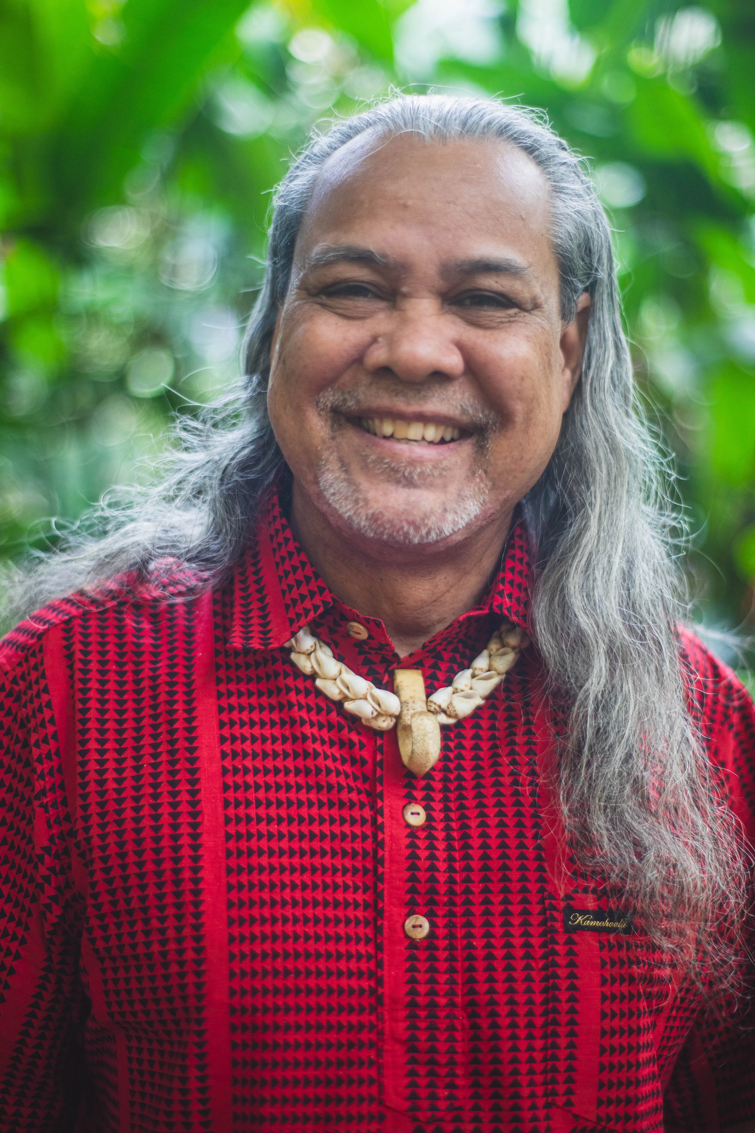 A smiling man with long gray hair and a mustache, wearing a red and black patterned shirt, with a shell necklace, outdoors with a green, leafy background.