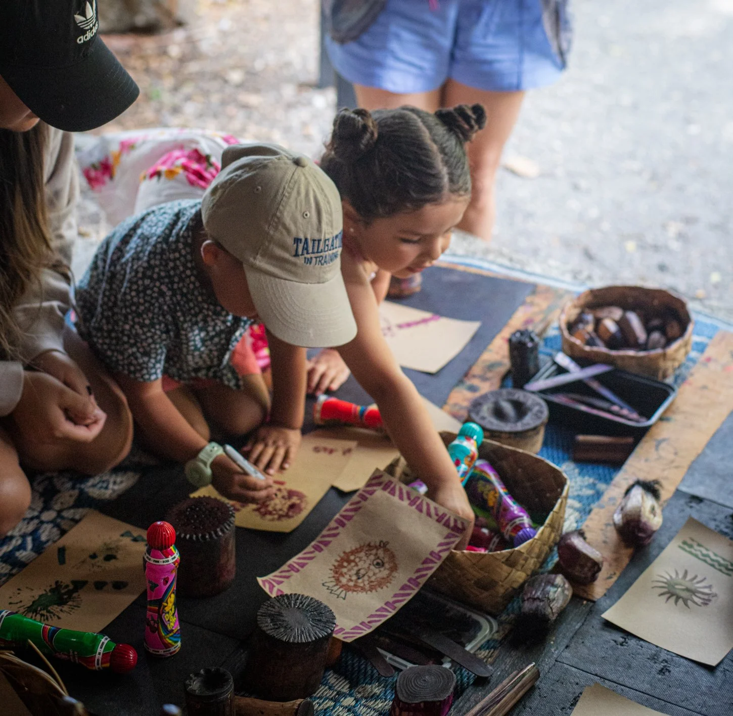 Children at an outdoor art station, coloring and drawing on paper with various art supplies including crayons, markers, and paper, surrounded by natural elements and decorative items.