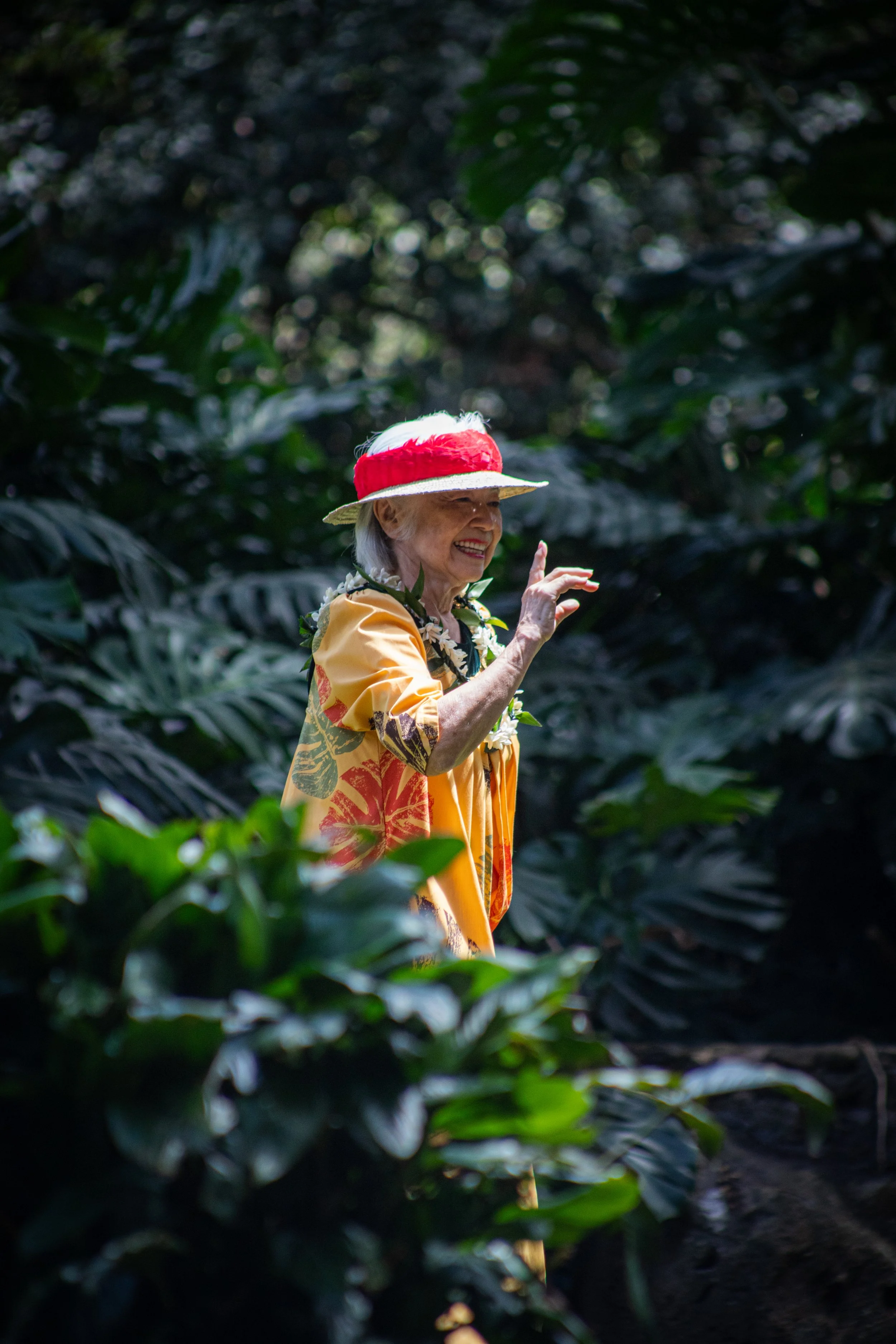 An elderly woman smiling and wearing a yellow dress, a floral lei, and a straw hat with a red band, walking through a lush, green forest.