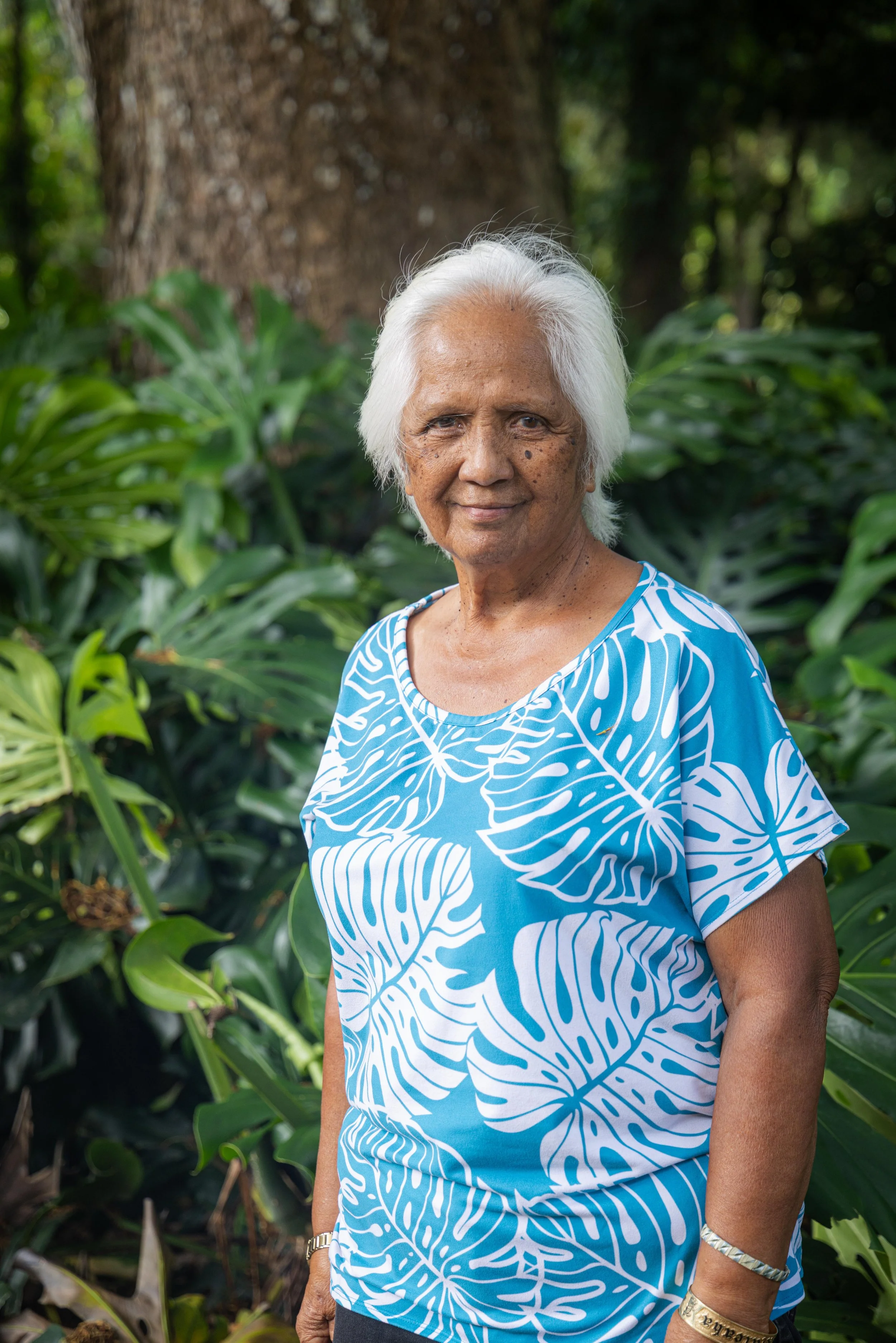 Elderly woman with white hair wearing a blue and white patterned dress, standing outdoors among green foliage and trees.