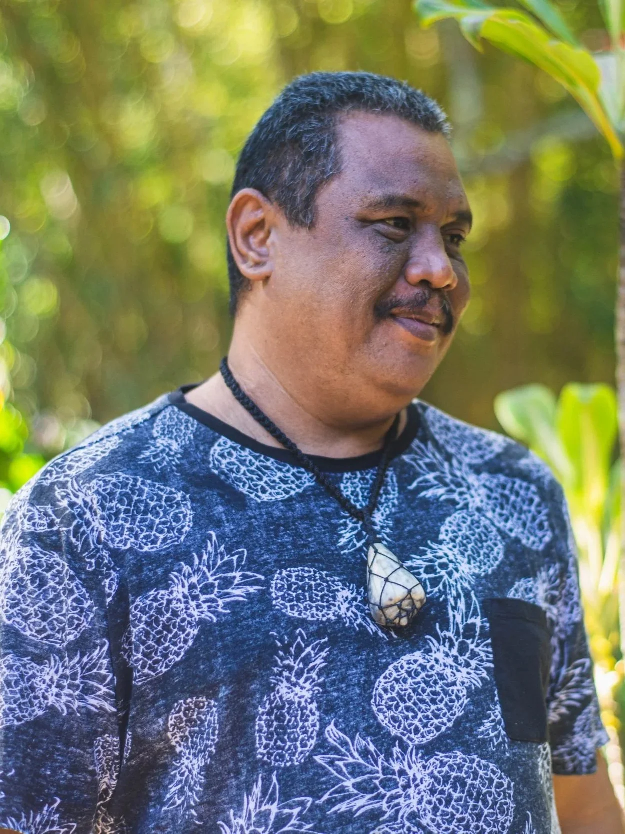 A middle-aged Polynesian man with short black hair and facial hair, wearing a patterned shirt with white floral designs on a dark background and a shell necklace, outdoors with blurred green foliage.