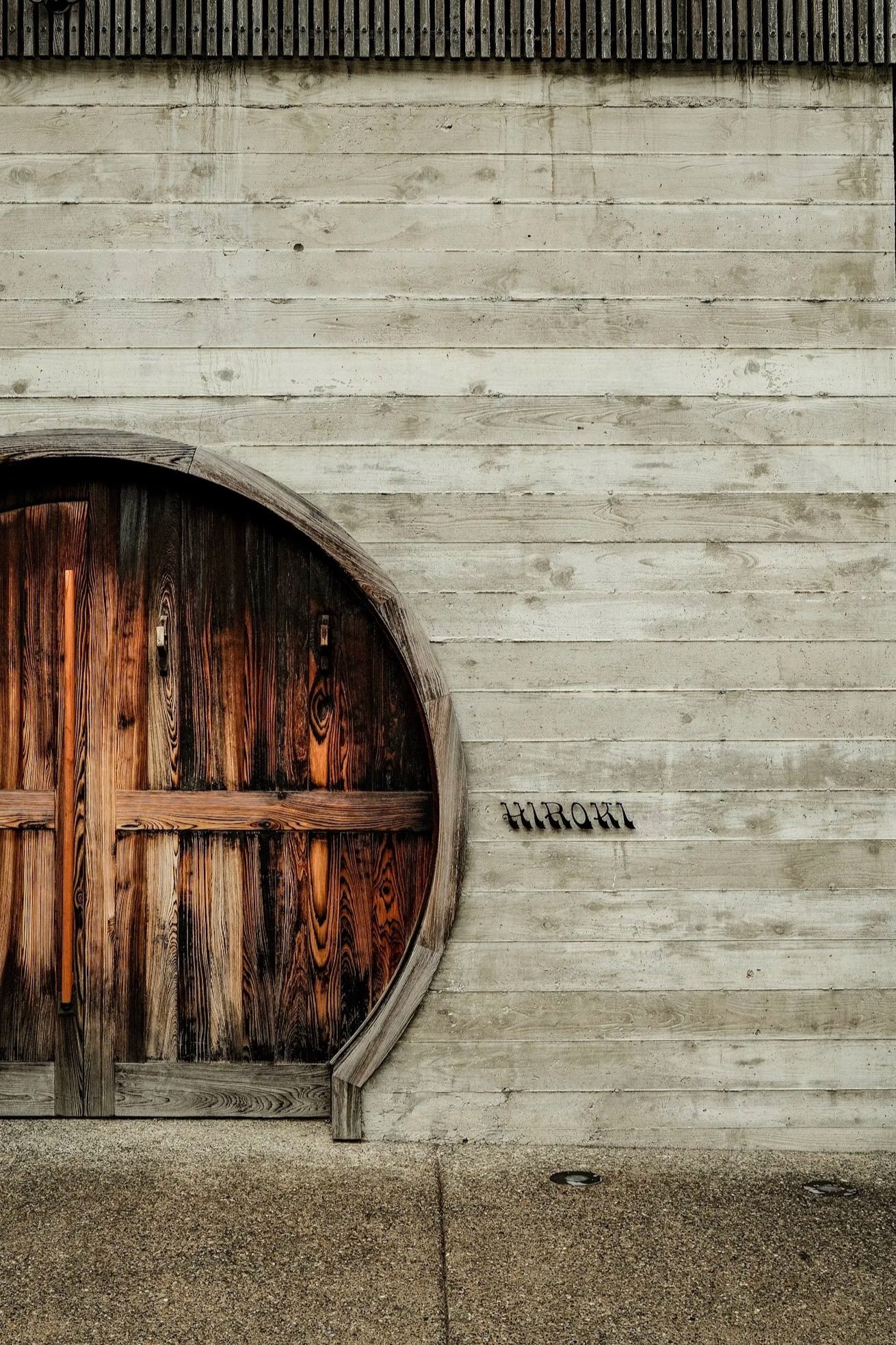 Wooden circular door with an arched top, set in a white weathered wooden wall. The door has vertical wooden planks with a natural finish. Below the door, there is a section of gravel and two small circular lights embedded in the ground, with the word 'HIROKI' written on the wall.