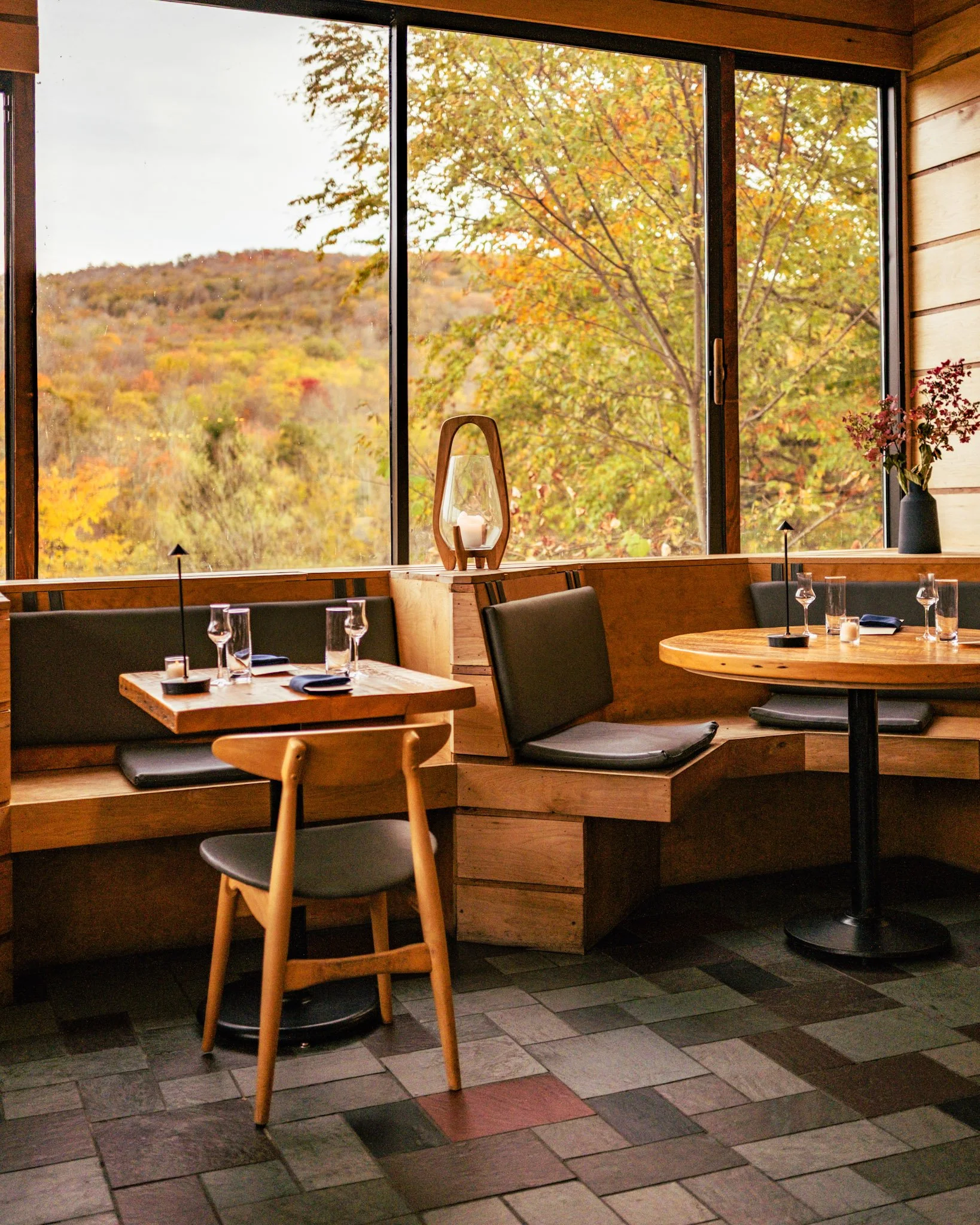 Interior of a restaurant with a wooden table and chairs, large windows showing a view of trees with autumn foliage, a candle in a wooden holder, a black vase with flowers, and a tiled floor.