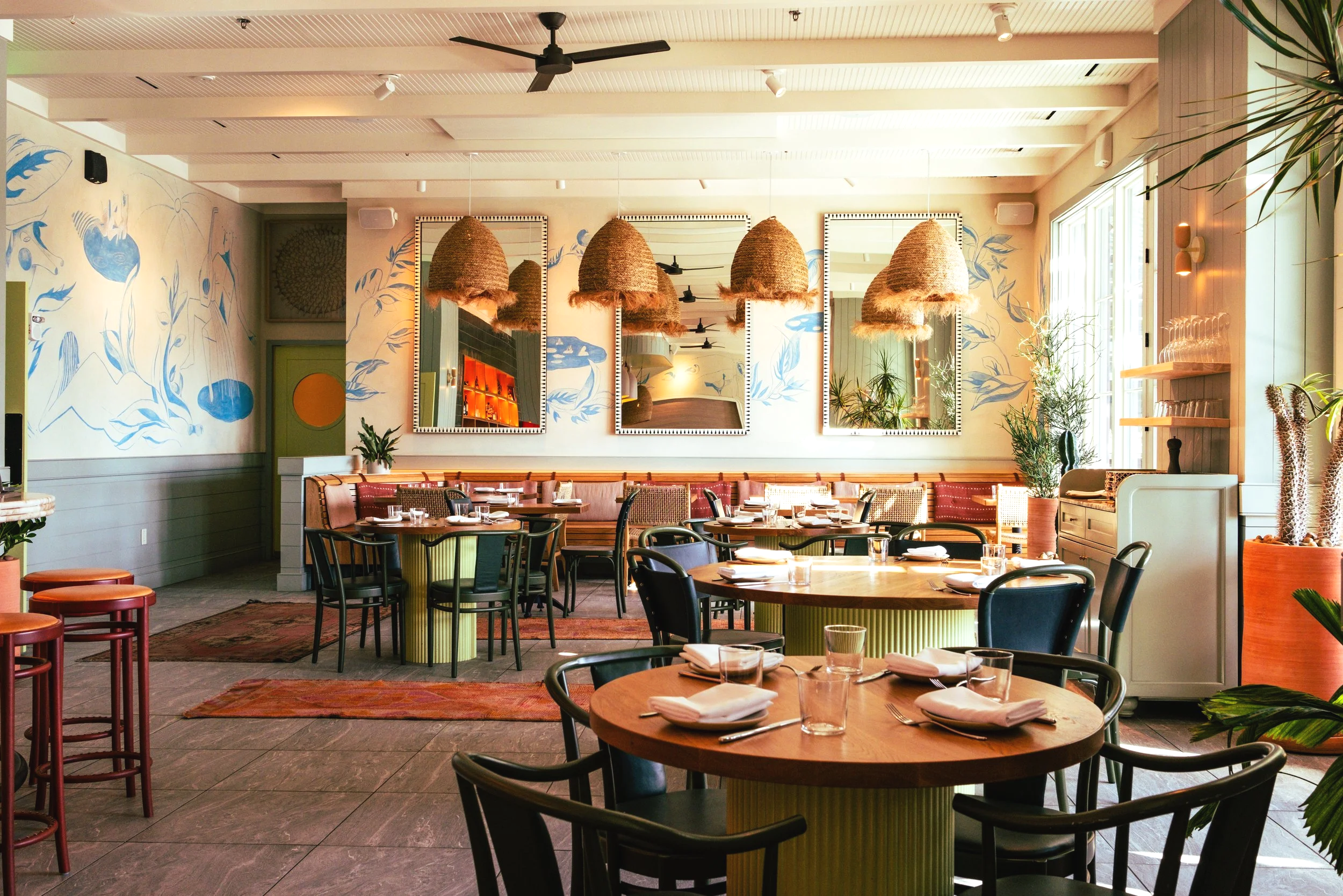 Interior of a modern restaurant with round and rectangular tables set with tableware, wooden and black chairs, wall art, large mirrors, hanging woven light fixtures, and lush green plants near large glass windows.