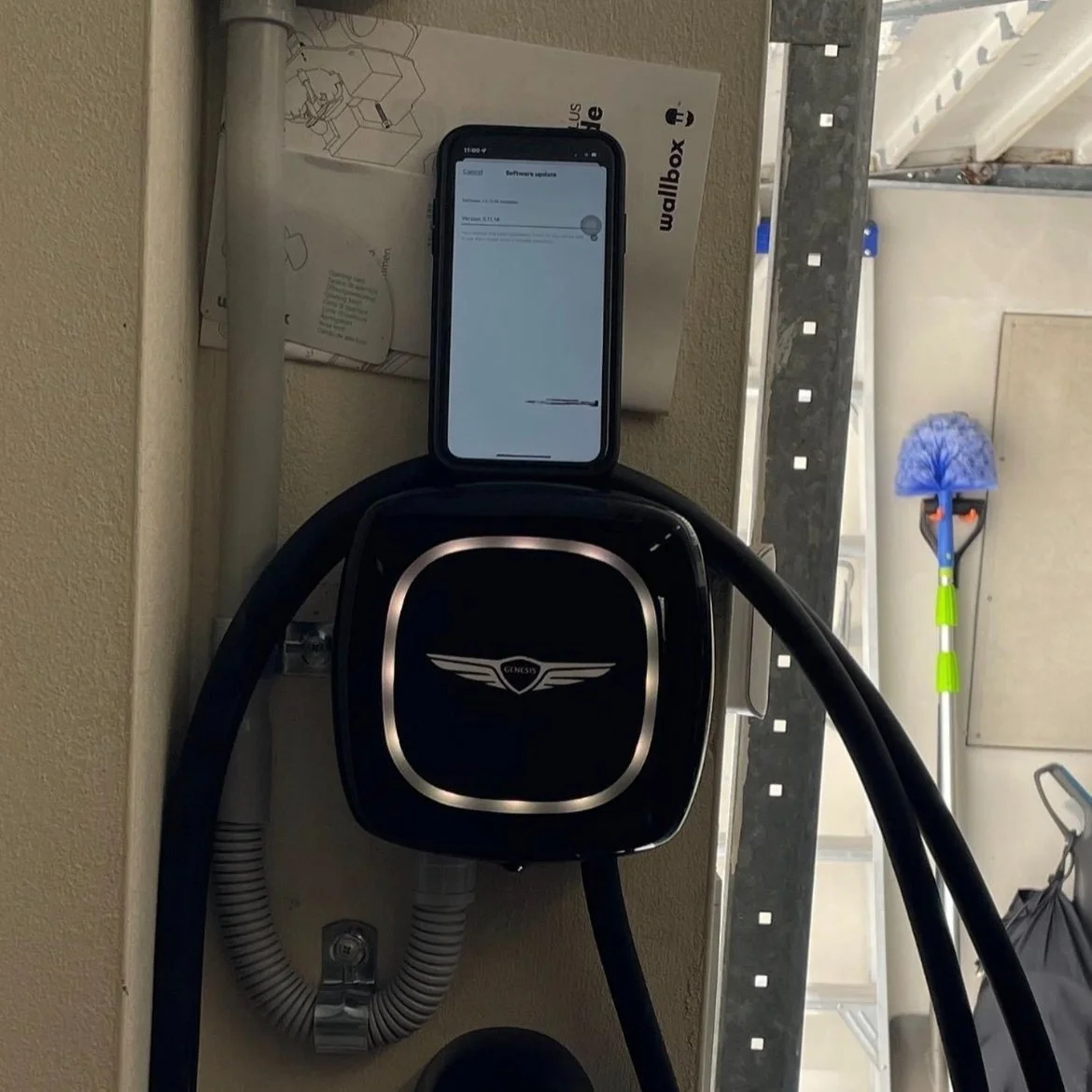 An electric car charging station with a Genesis logo, a smartphone placed on top showing a software update screen, a wallbox user manual nearby, and a dustpan and broom in the background in a garage.