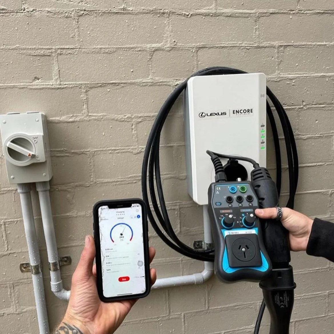 Person holding an electric vehicle charging plug, connected to a charging station on a brick wall, and monitoring the charging progress on a smartphone.