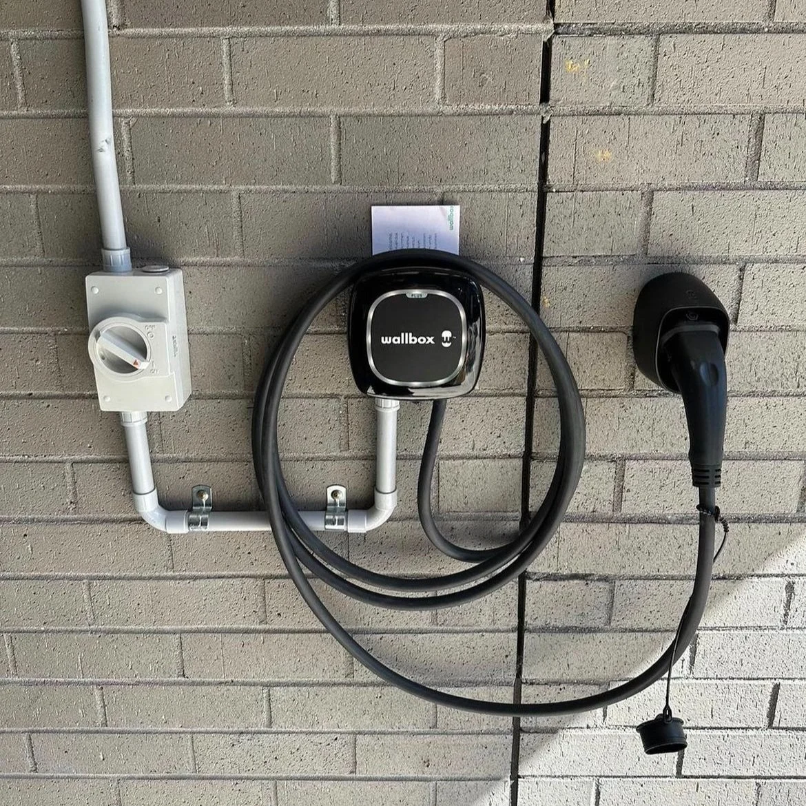 Electric vehicle charging station setup on a brick wall, including a power socket, a Wallbox charger, and an electric plug.