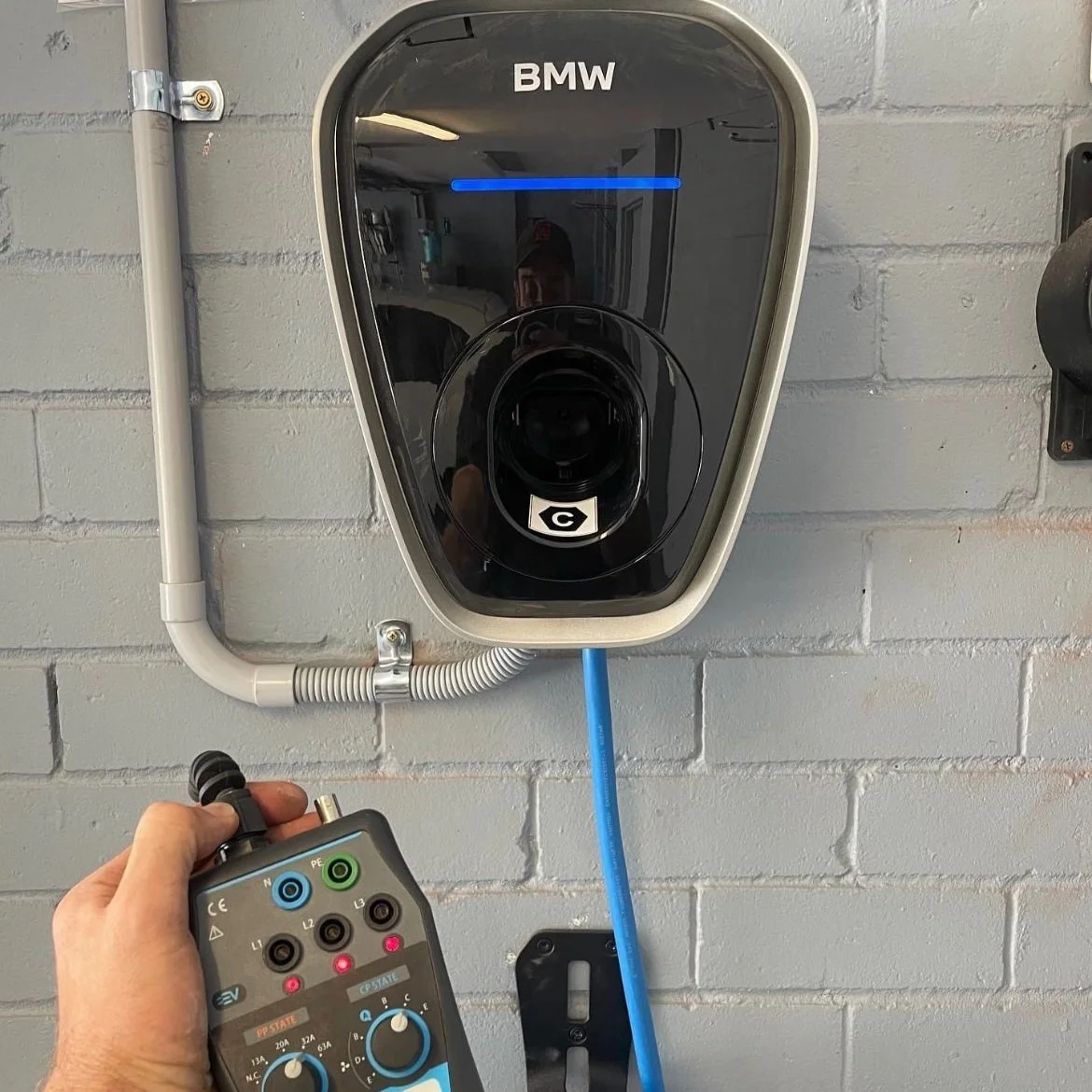 An electric vehicle charging station labeled BMW with a person holding a testing device attached to a blue charging cable.