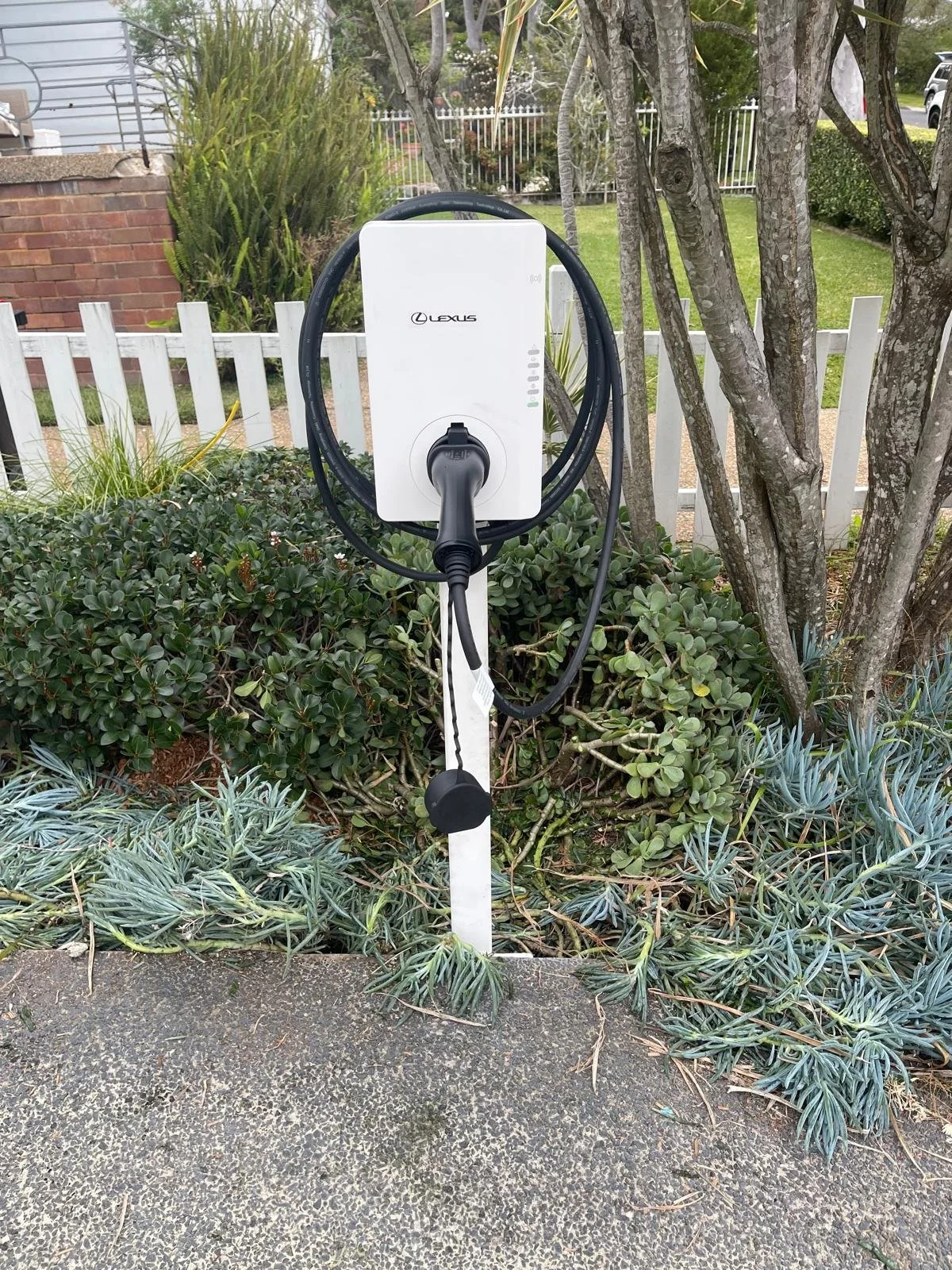 Electric vehicle charging station outdoors surrounded by plants and trees, with a white picket fence and residential area in the background.