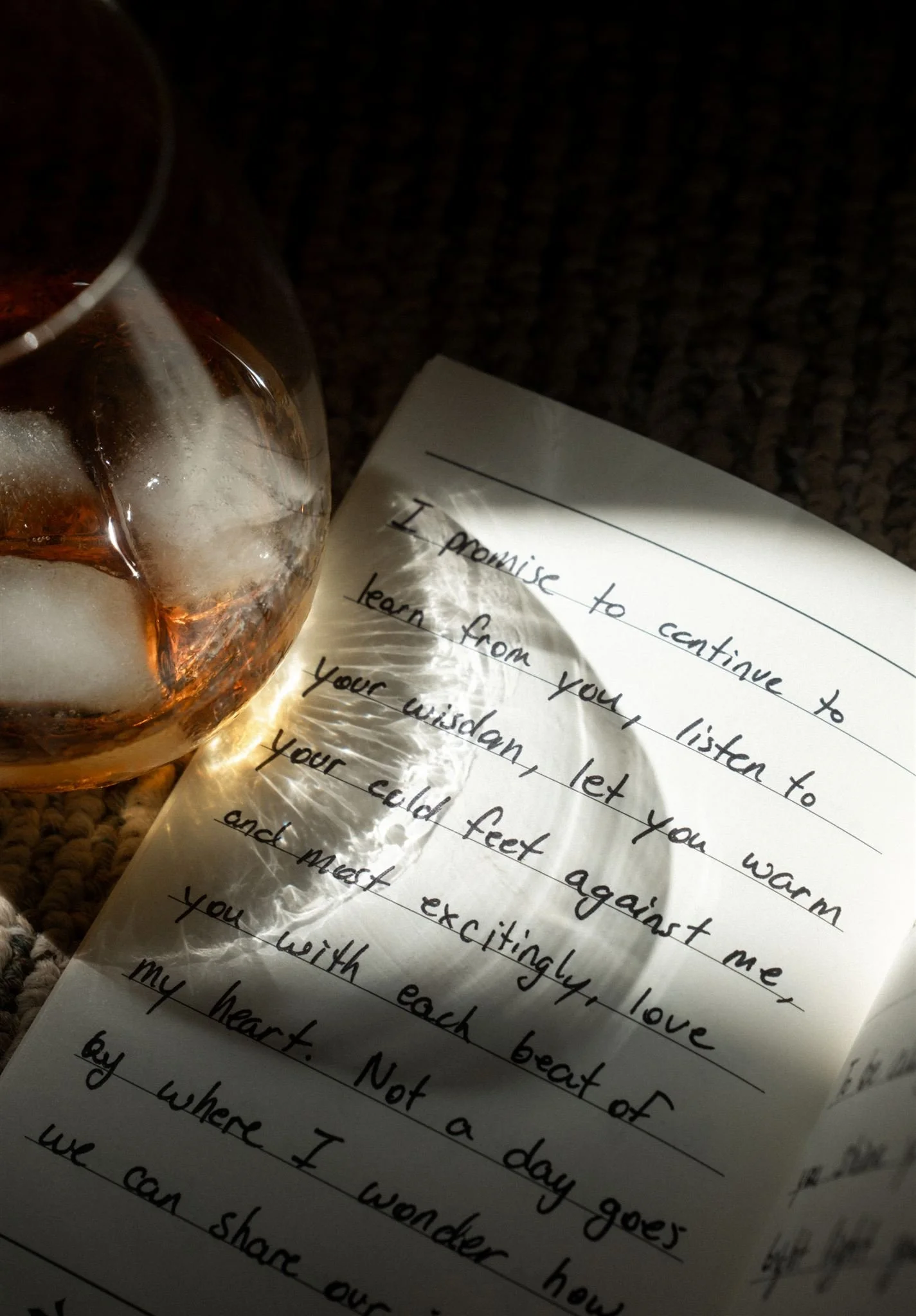 A handwritten note on lined paper with a glass of whiskey with ice, casting a reflection of the note. The note contains a promise to continue learning and loving.