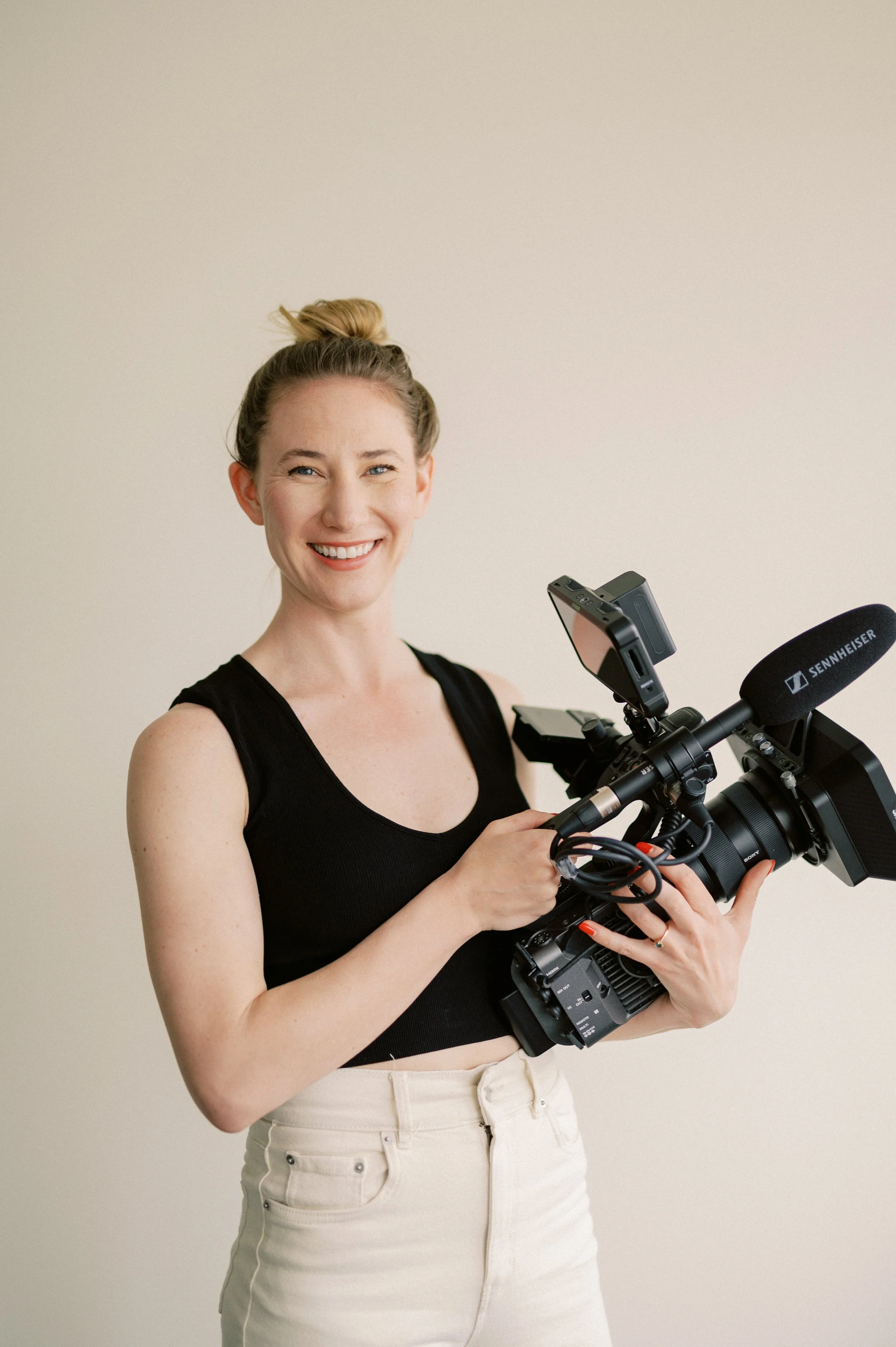 A smiling woman with blonde hair tied in a bun, wearing a black sleeveless top and beige pants, holding a professional video camera with a mounted microphone against a plain light-colored wall.