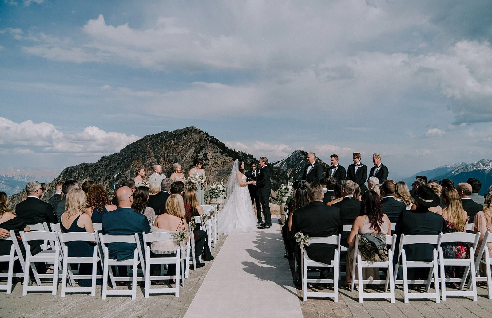 Outdoor wedding ceremony on a mountain with a bride and groom exchanging vows, surrounded by seated guests, mountain peaks in the background, under a cloudy sky.