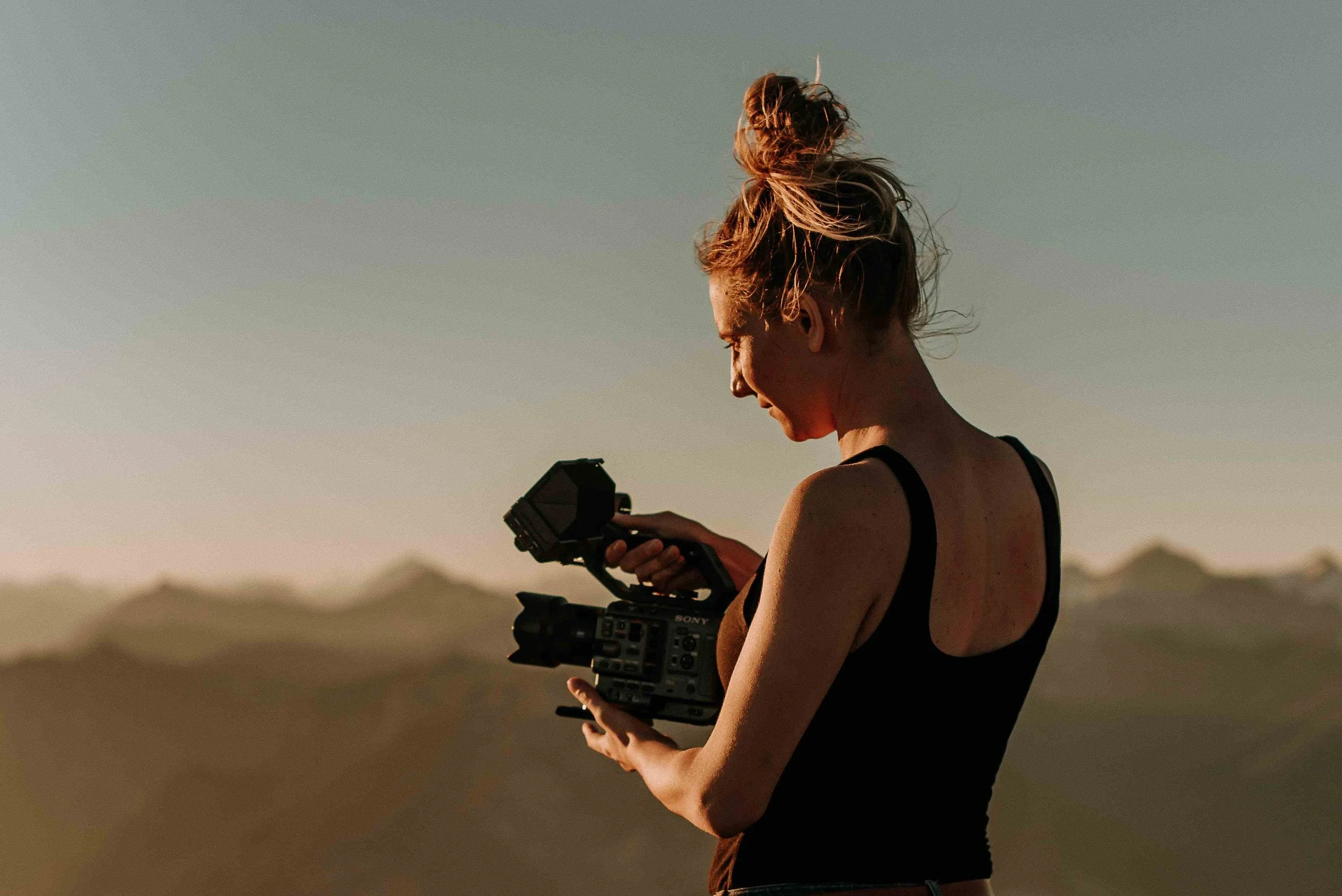 A woman with a messy bun holding a video camera outdoors in a desert landscape during sunset.