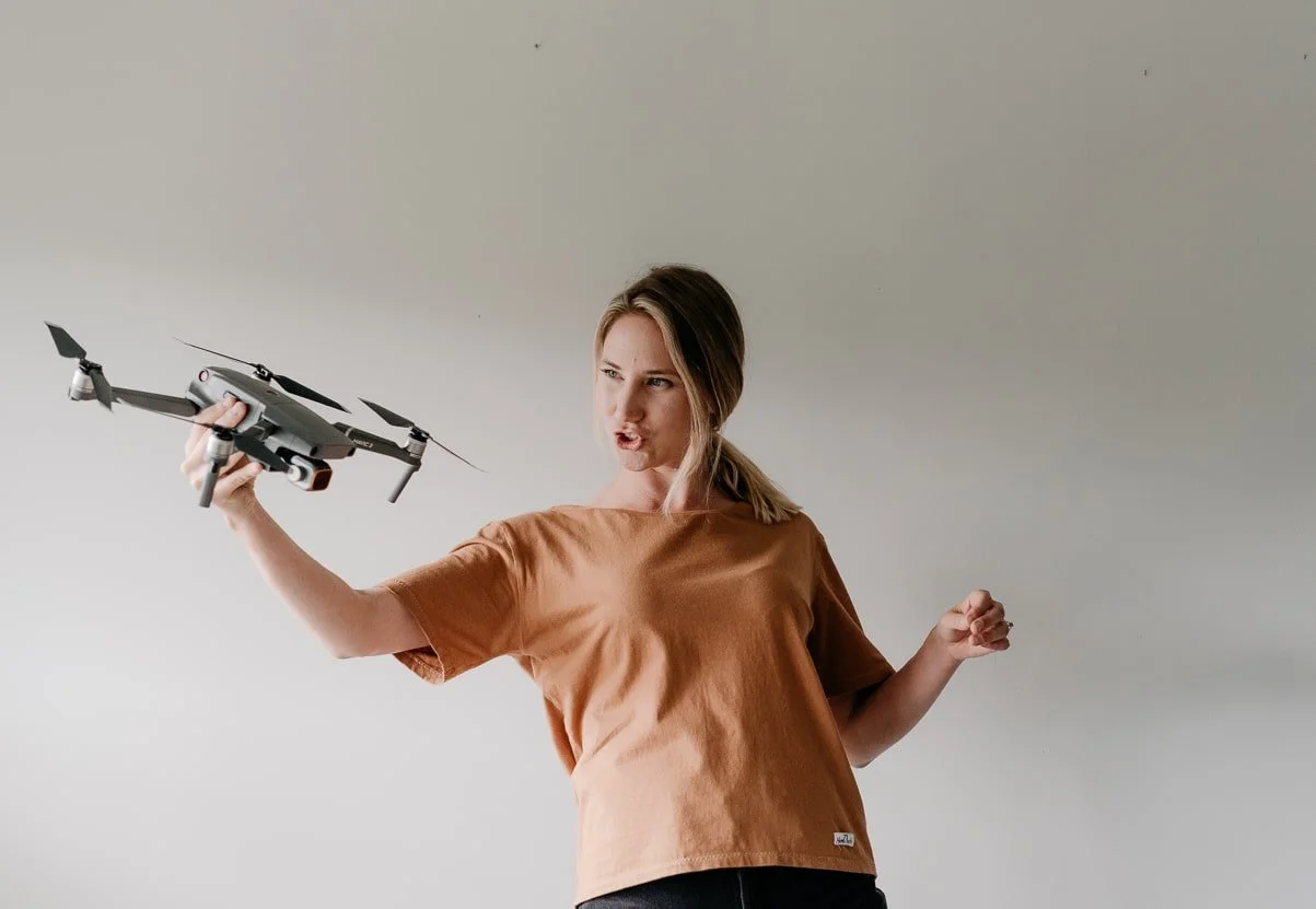 A young woman with blonde hair in a ponytail, wearing an orange T-shirt, holding a grey drone and making a fierce expression.