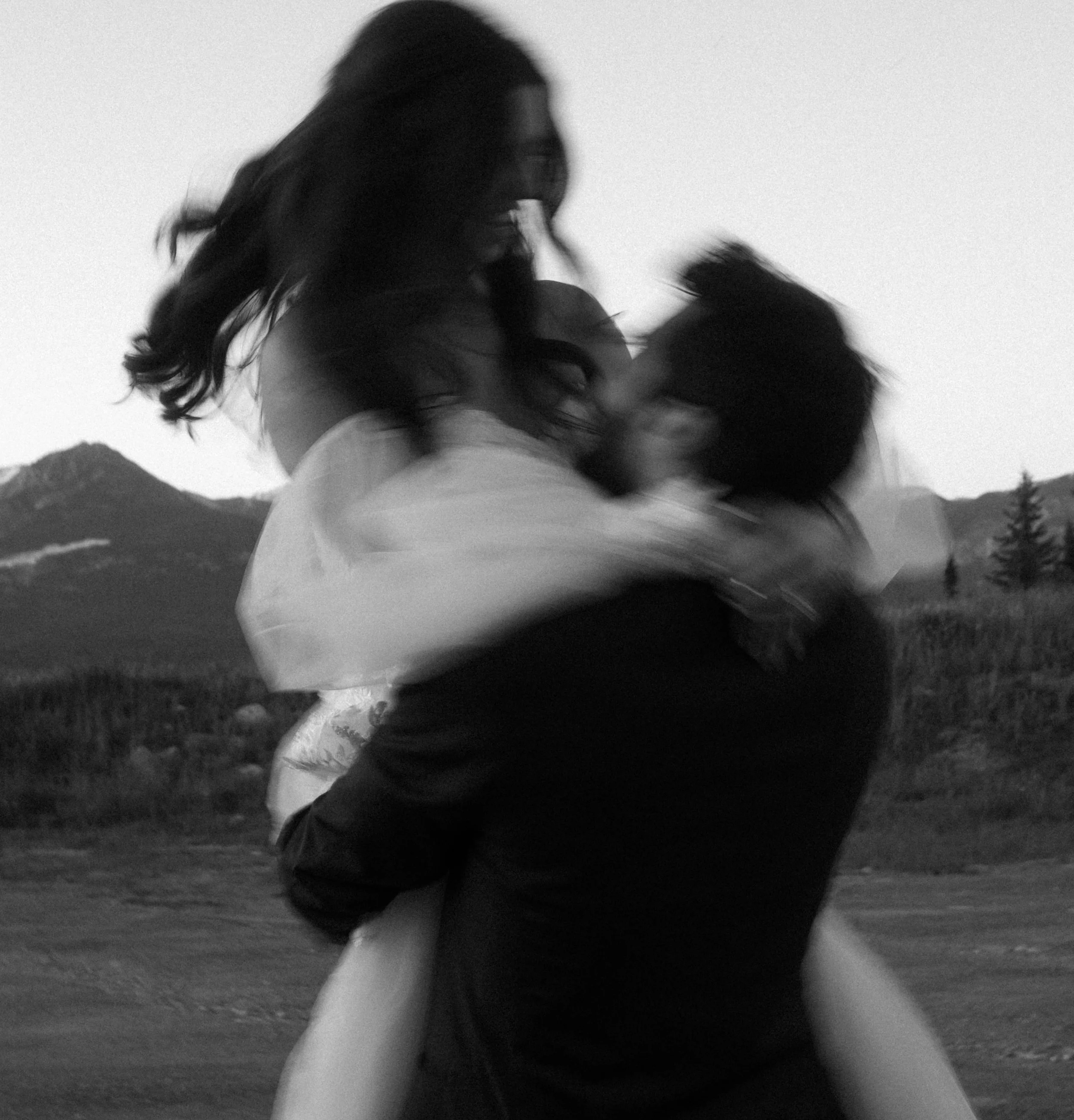A black and white photo of a couple embracing and kissing outdoors, with mountains and trees in the background.