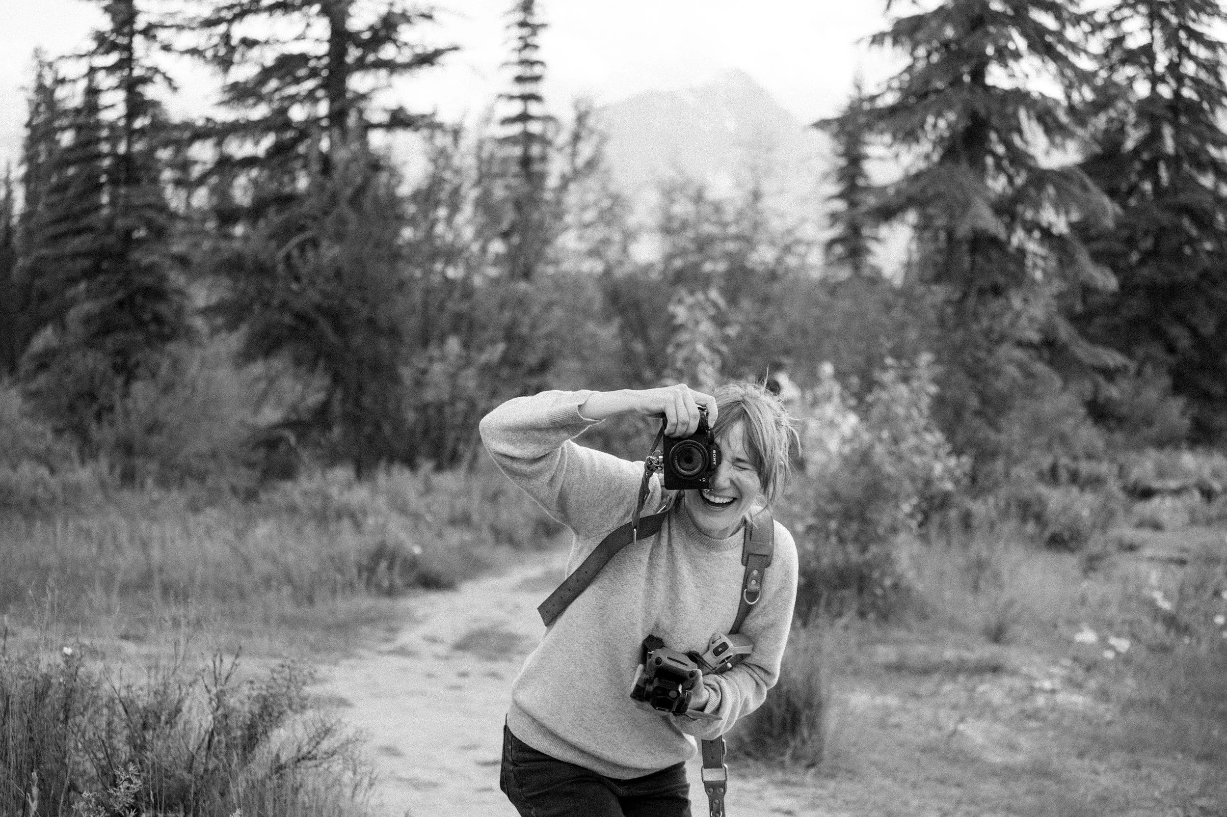 A smiling woman with a camera and a backpack, outdoors in a forested area with trees and mountains in the background.