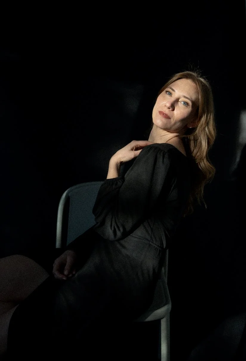 A woman with light skin and wavy brown hair sitting on a chair against a dark background, looking at the camera with a neutral expression.