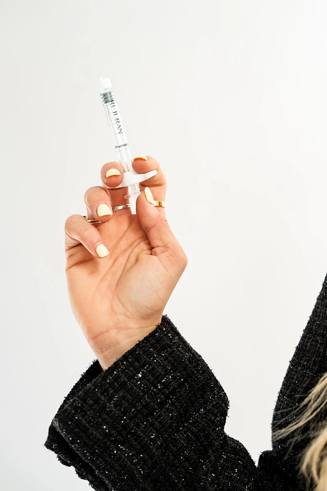 Close-up of a person's hand holding a small syringe with a white cap and clear liquid, against a plain white background. The person is wearing a black, textured, long-sleeve garment and has manicured nails with light-colored polish.