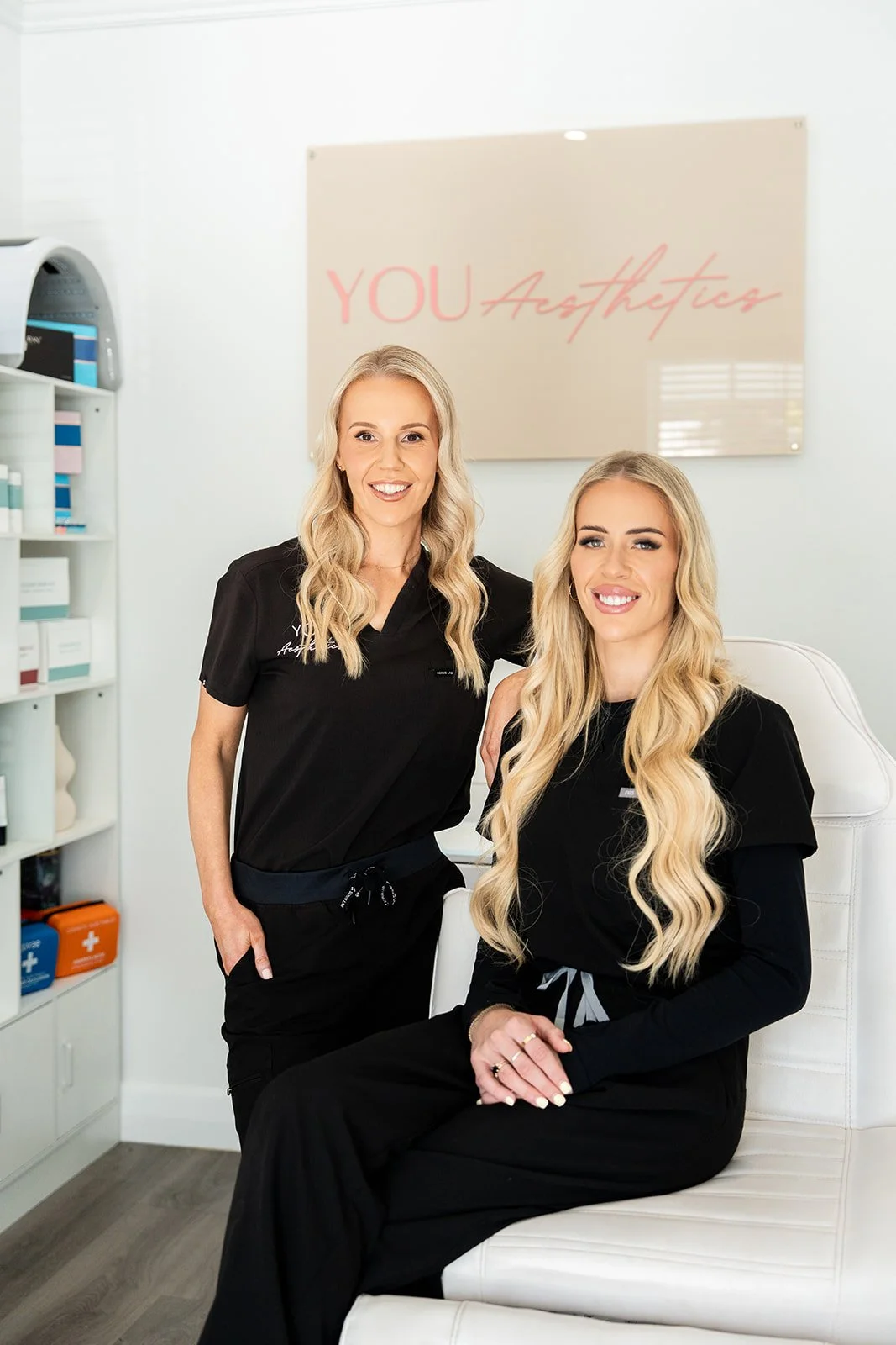 Two women with long blonde hair smiling in a spa or aesthetic clinic with a sign behind them reading 'You Aesthetics'. One is standing, and the other is sitting in a white chair.