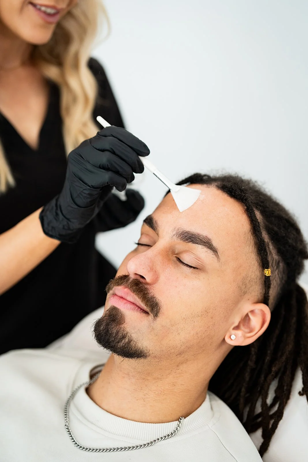 A woman with blonde hair, wearing black gloves, is applying a treatment to a man's forehead with a small brush. The man has closed eyes, a mustache, a beard, and dreadlocks, and is lying down with a relaxed expression.