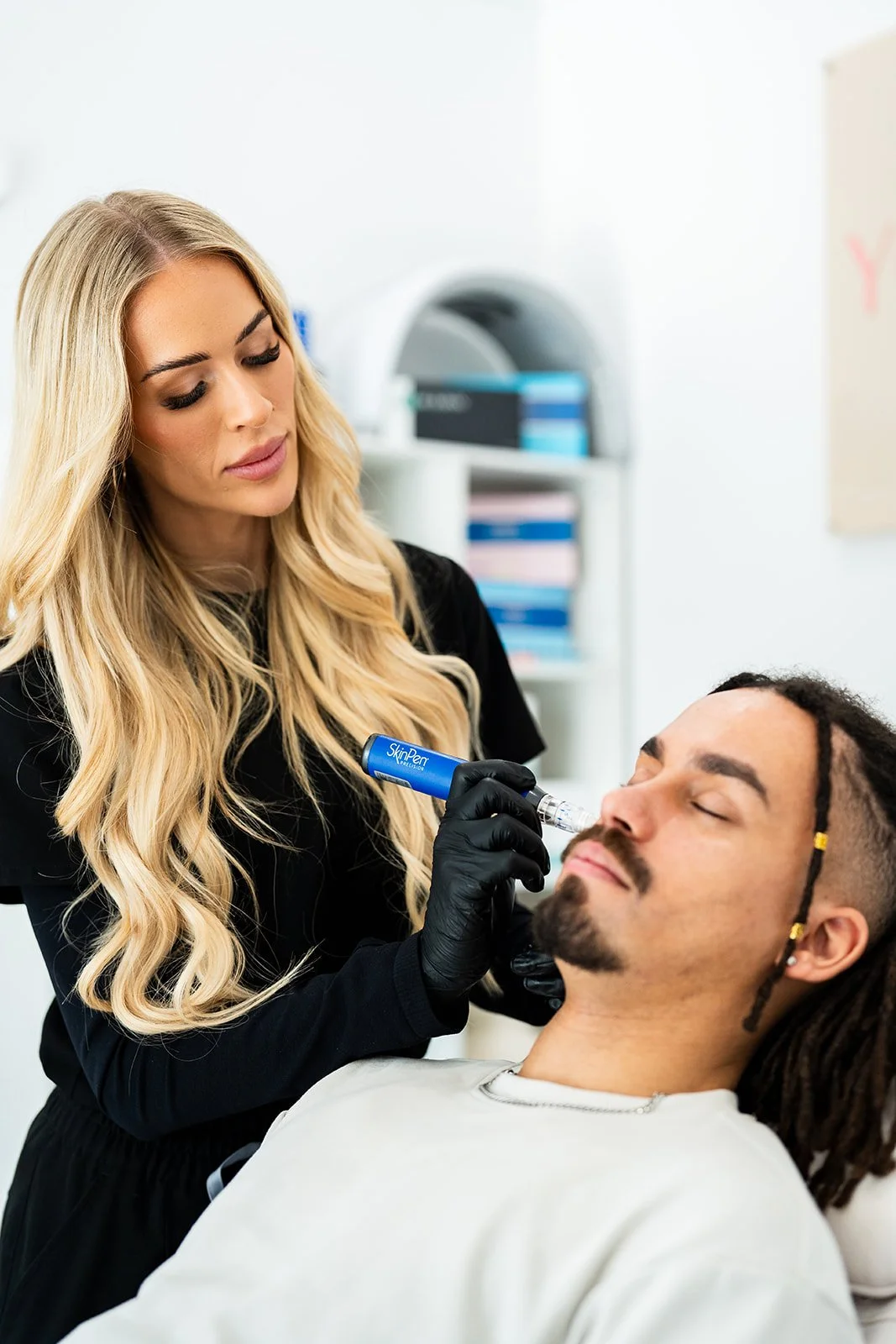 A woman with long blonde hair and black gloves is holding a SkinPen microneedling device to a man's face while he is reclined with eyes closed.