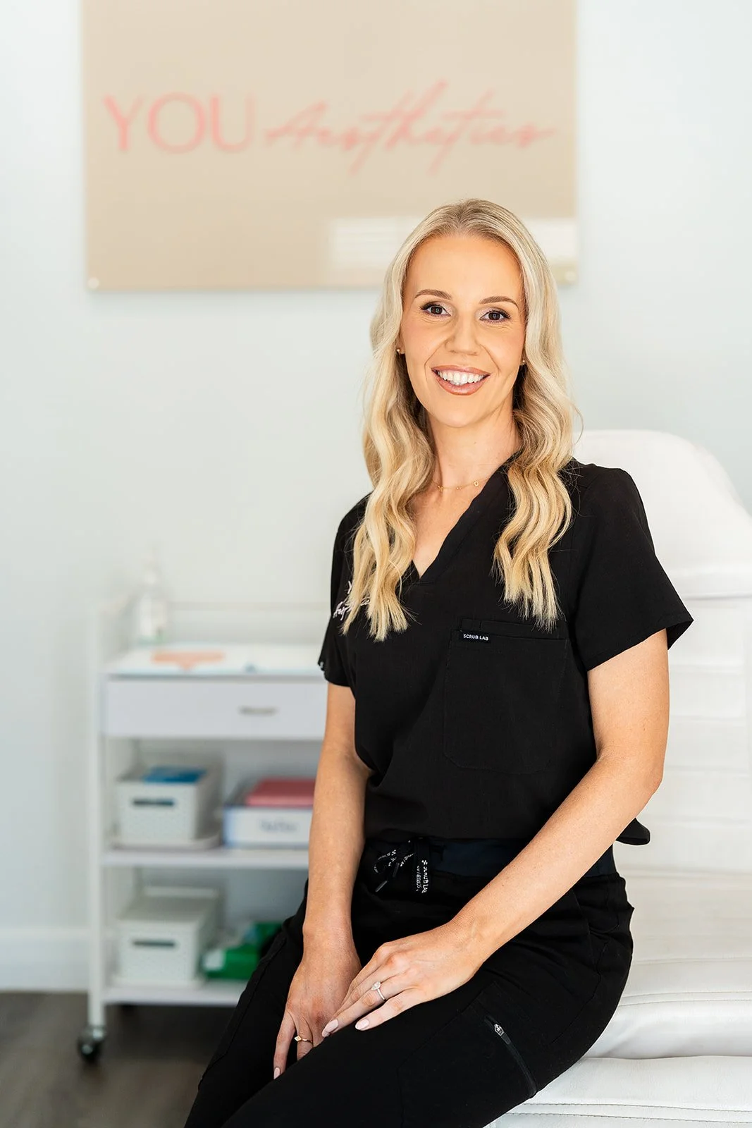 A smiling blonde woman in black scrubs sitting on a white chair in a medical office or clinic, with a sign that reads 'YOU Aesthetics' in pink in the background.